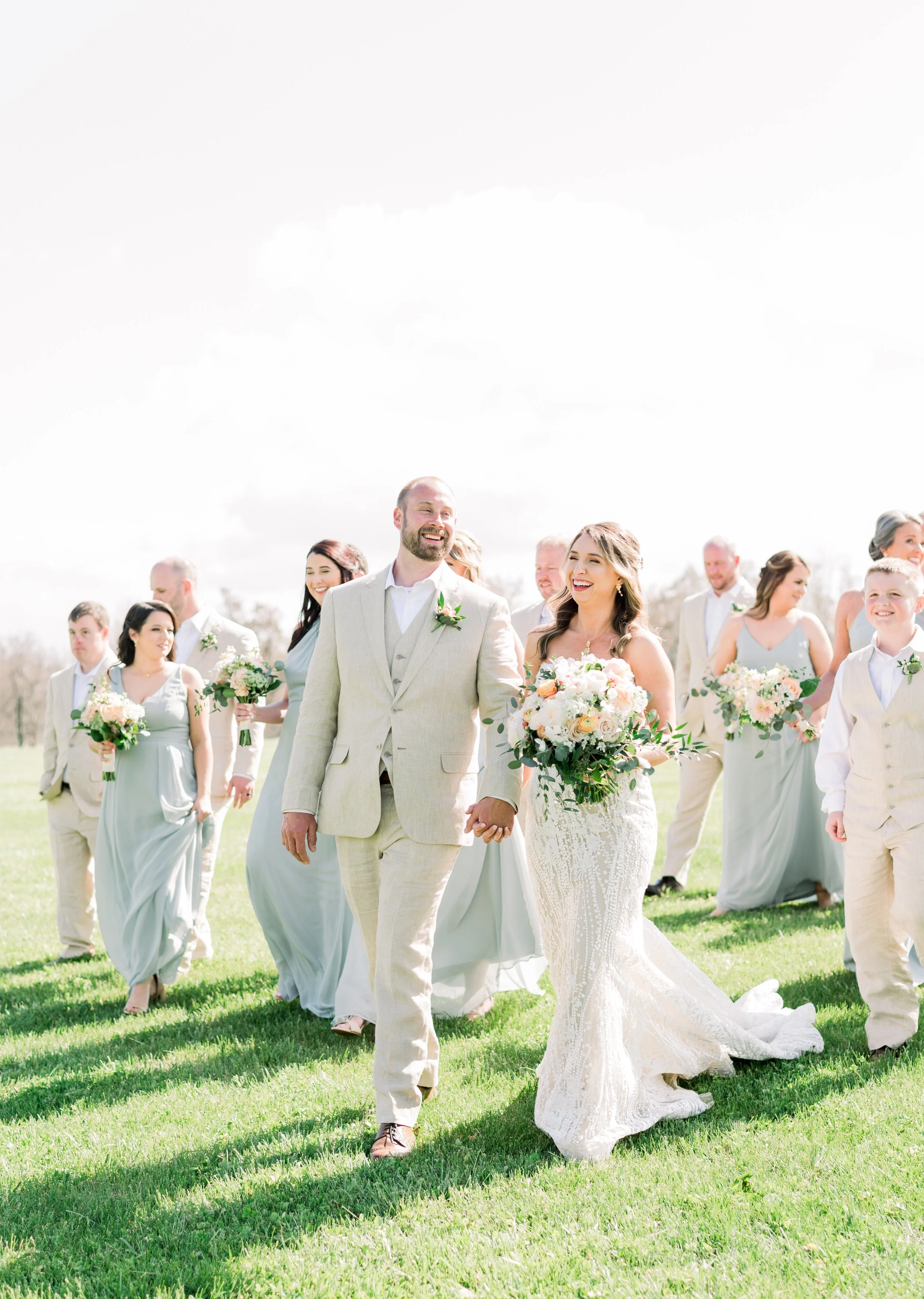 Bride and groom walking hand in hand with wedding party outdoors on green grass, smiling and holding bouquets, under bright sky.