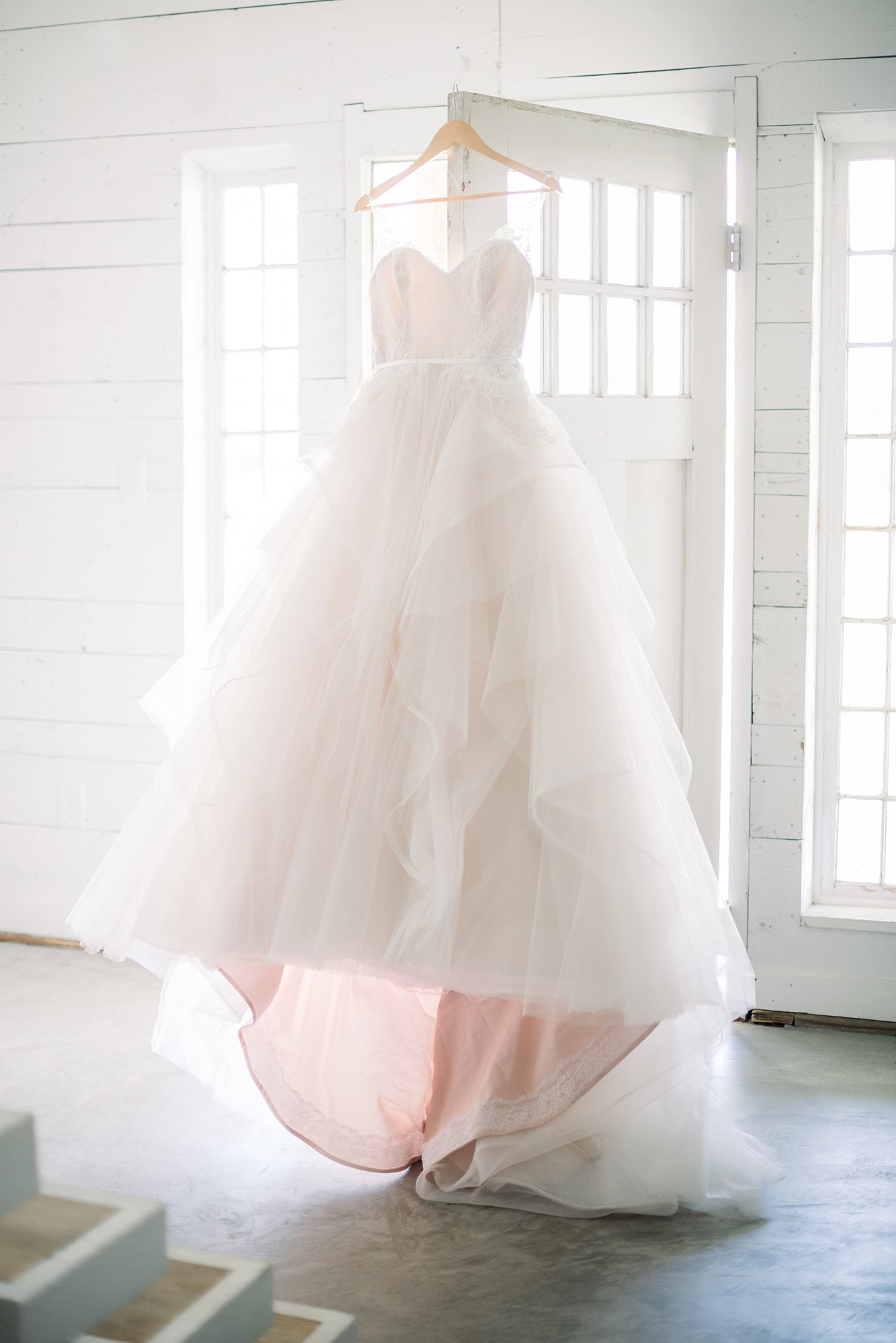A white wedding dress hanging on a wooden hanger in front of a window with sunlight coming through.
