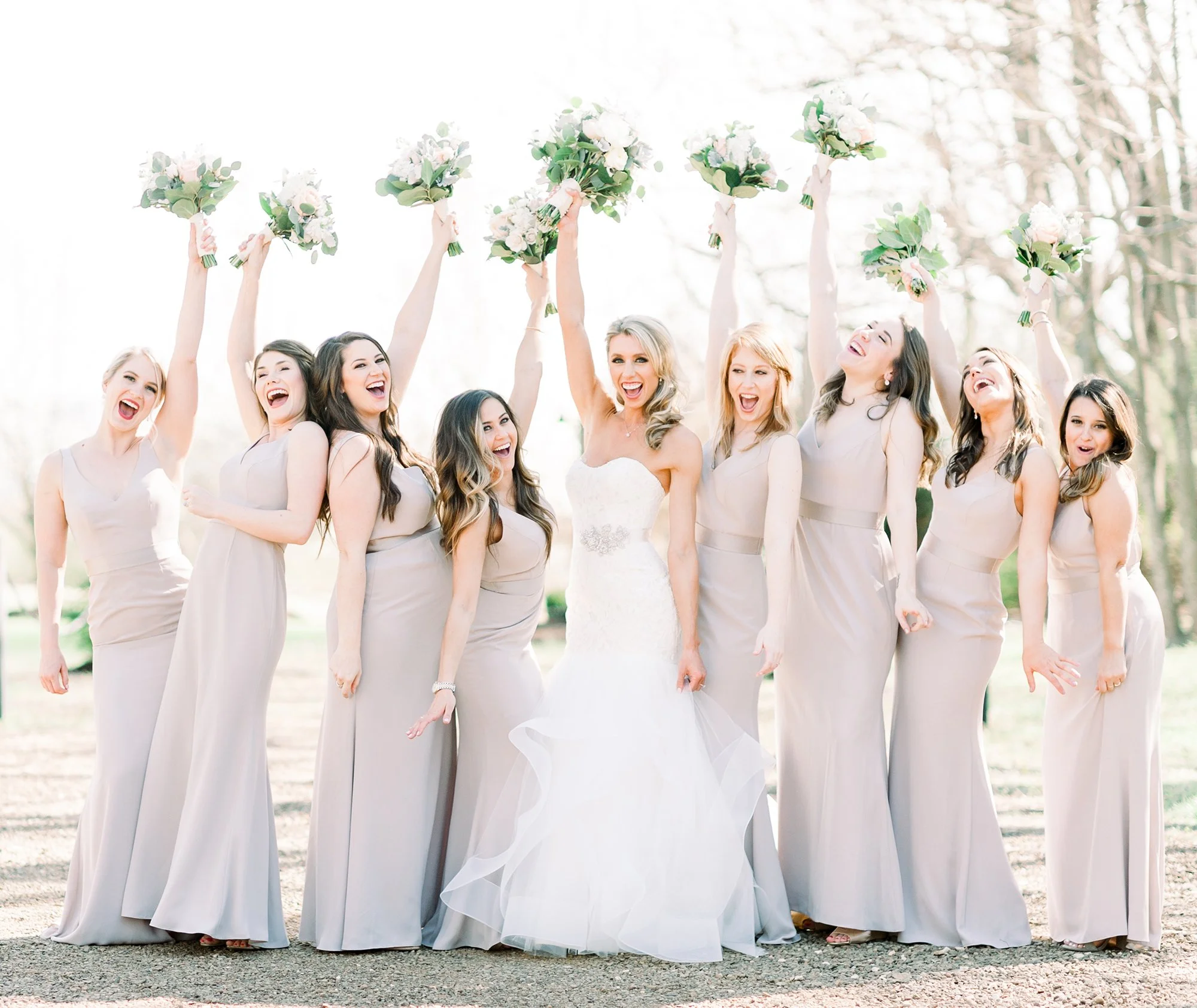 A bride and eight bridesmaids celebrating outdoors, holding bouquets and laughing, with the bride in the center wearing a white wedding dress and the bridesmaids in matching light-colored dresses.