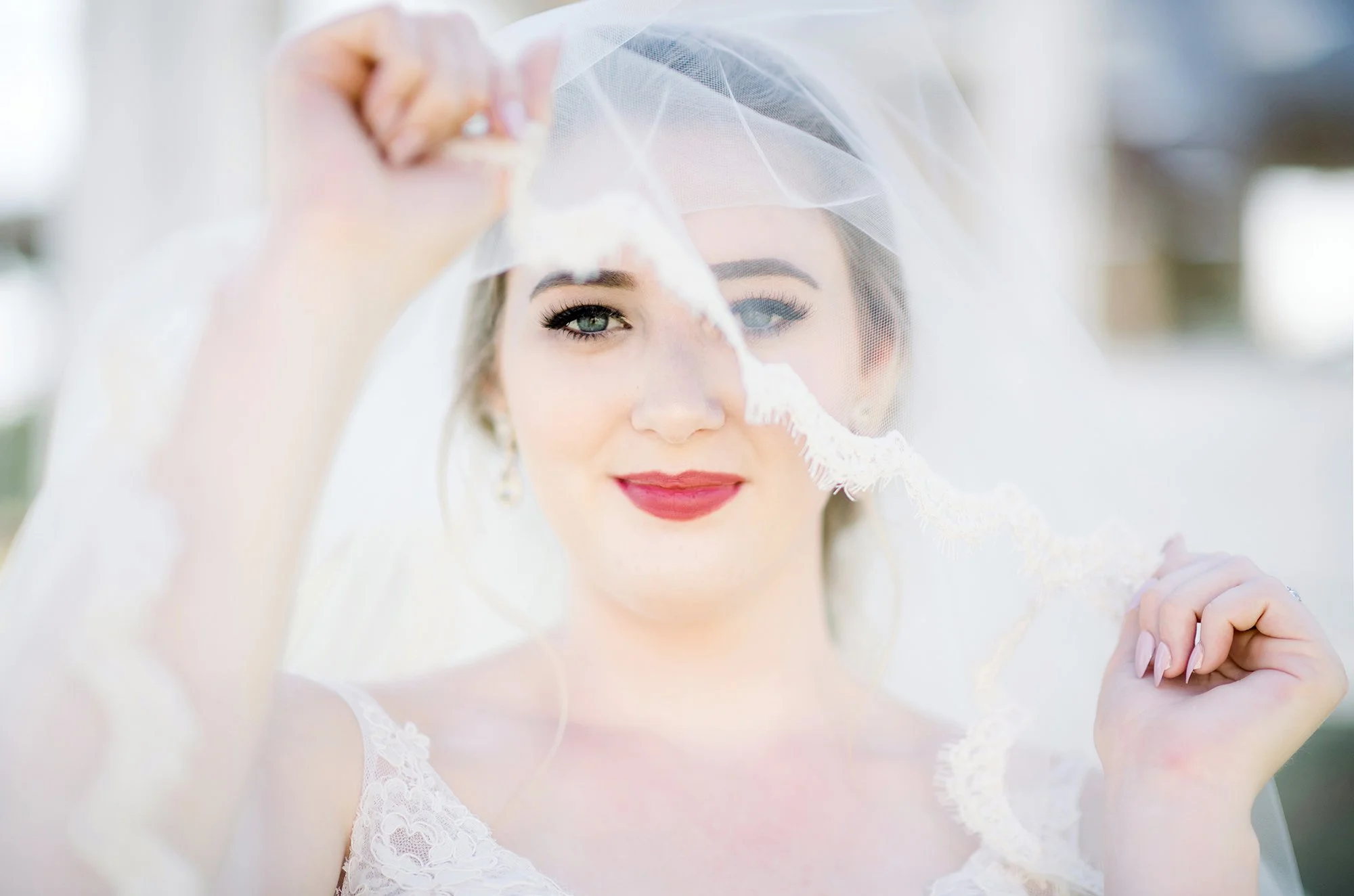 Close-up of a bride holding her wedding veil in front of her face, smiling softly, with visible makeup and earrings.