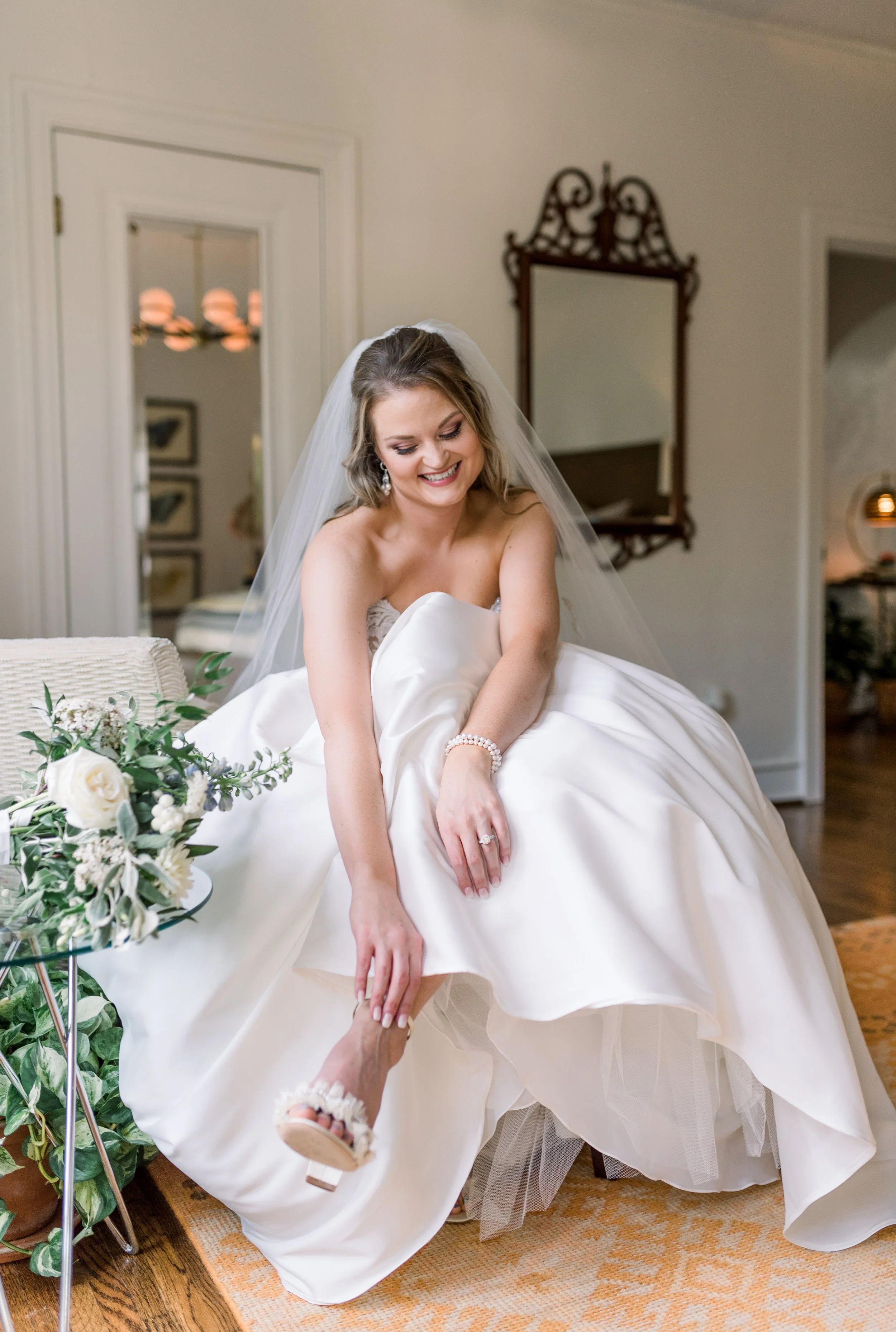 A bride getting dressed in a white wedding gown, smiling while putting on her shoe, with a bouquet on a stand nearby in a well-lit room.