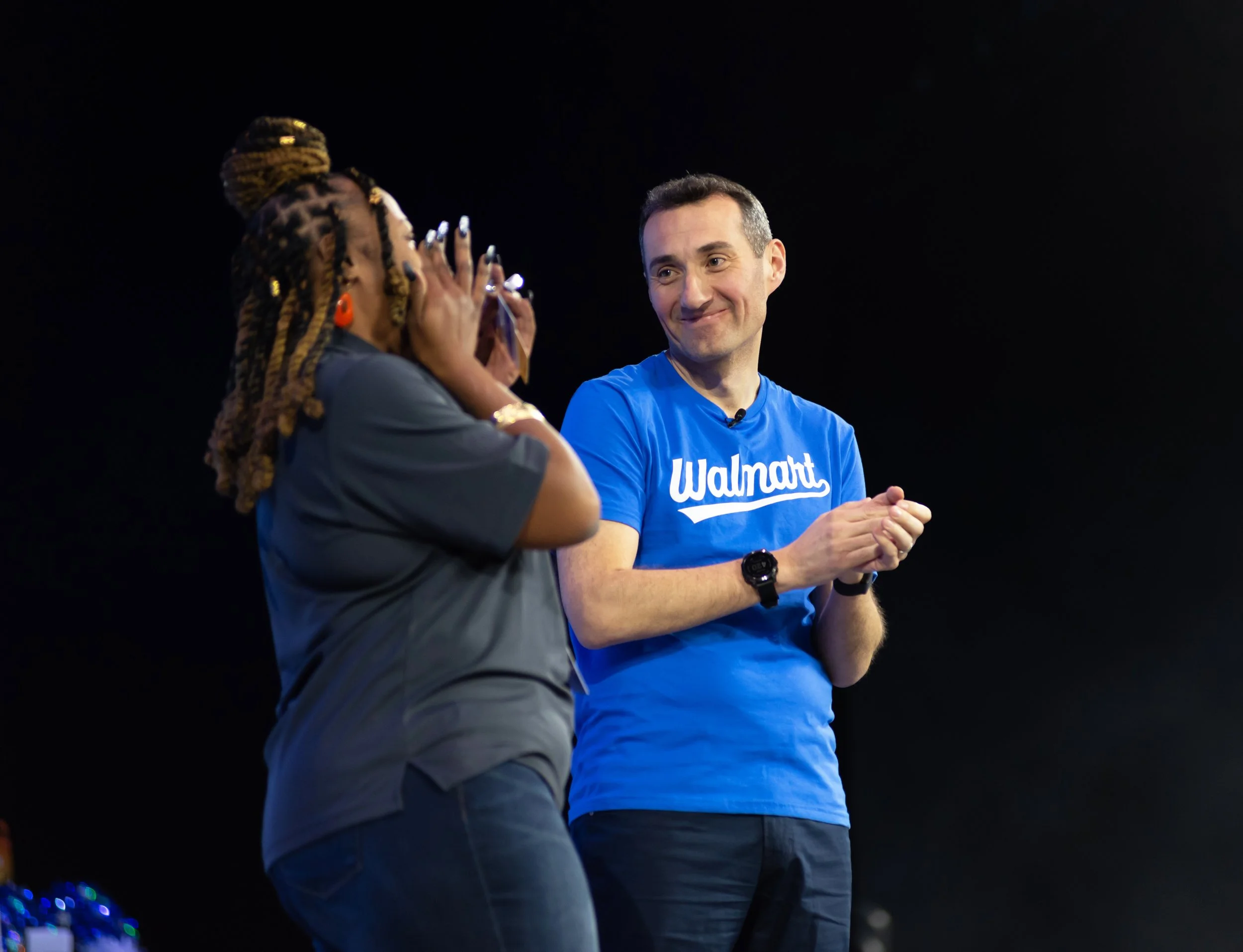 A woman with braided hair taking a photo or video of a man wearing a blue Walmart t-shirt, smiling on stage against a black background.