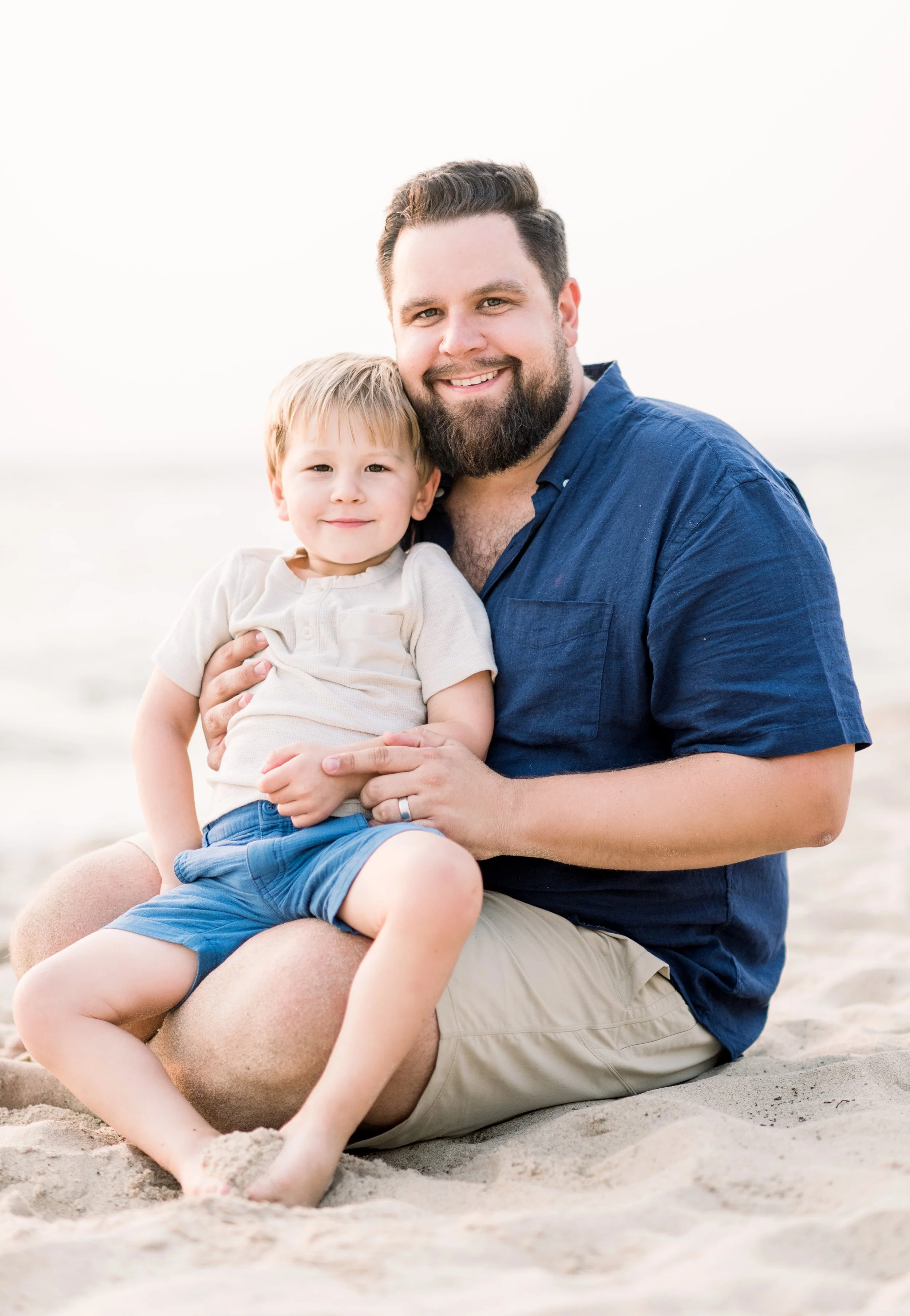 A man with a beard sitting on the sand at the beach, holding and smiling with a young boy, both looking at the camera.