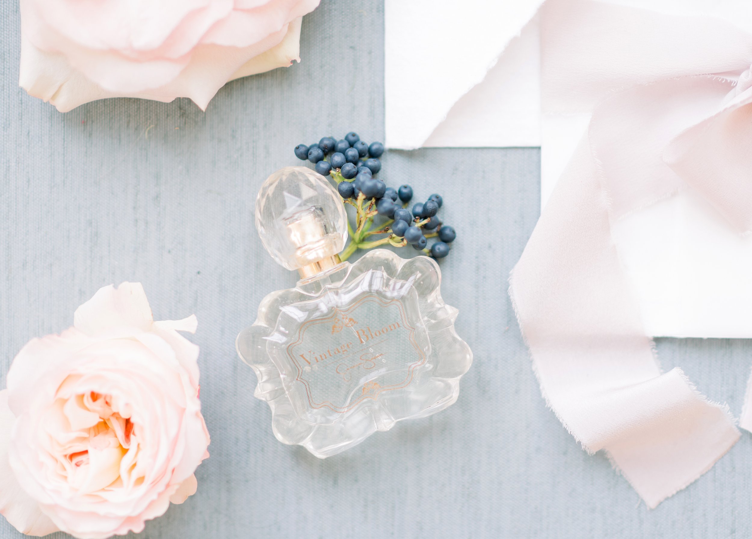 A vintage flower perfume bottle with pink label, surrounded by pink roses, a sprig of blue berries, soft pink ribbon, and torn white paper on a light blue background.
