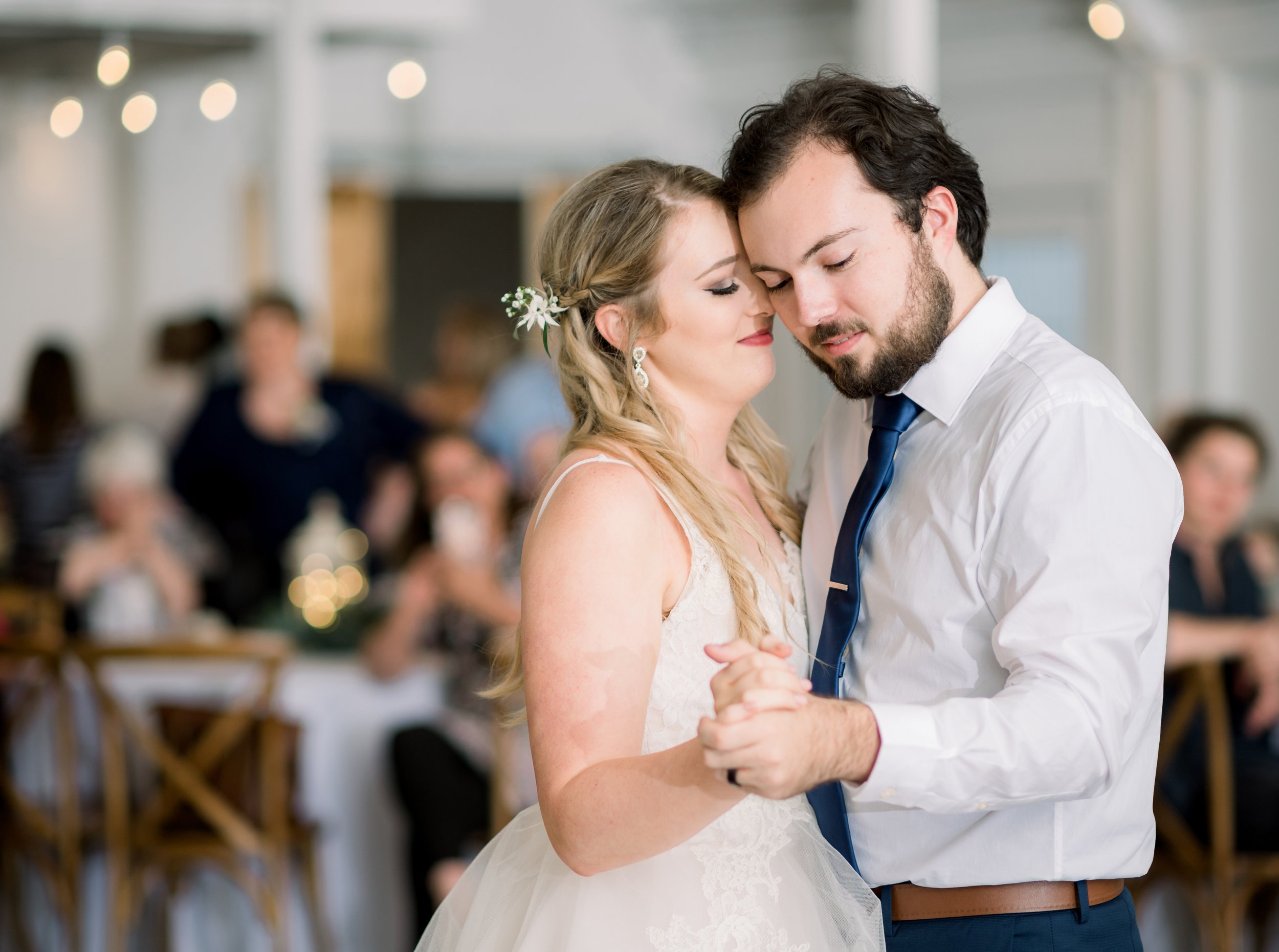 A bride and groom sharing a dance, eyes closed, at wedding reception; background guests seated at tables, blurred lighting.