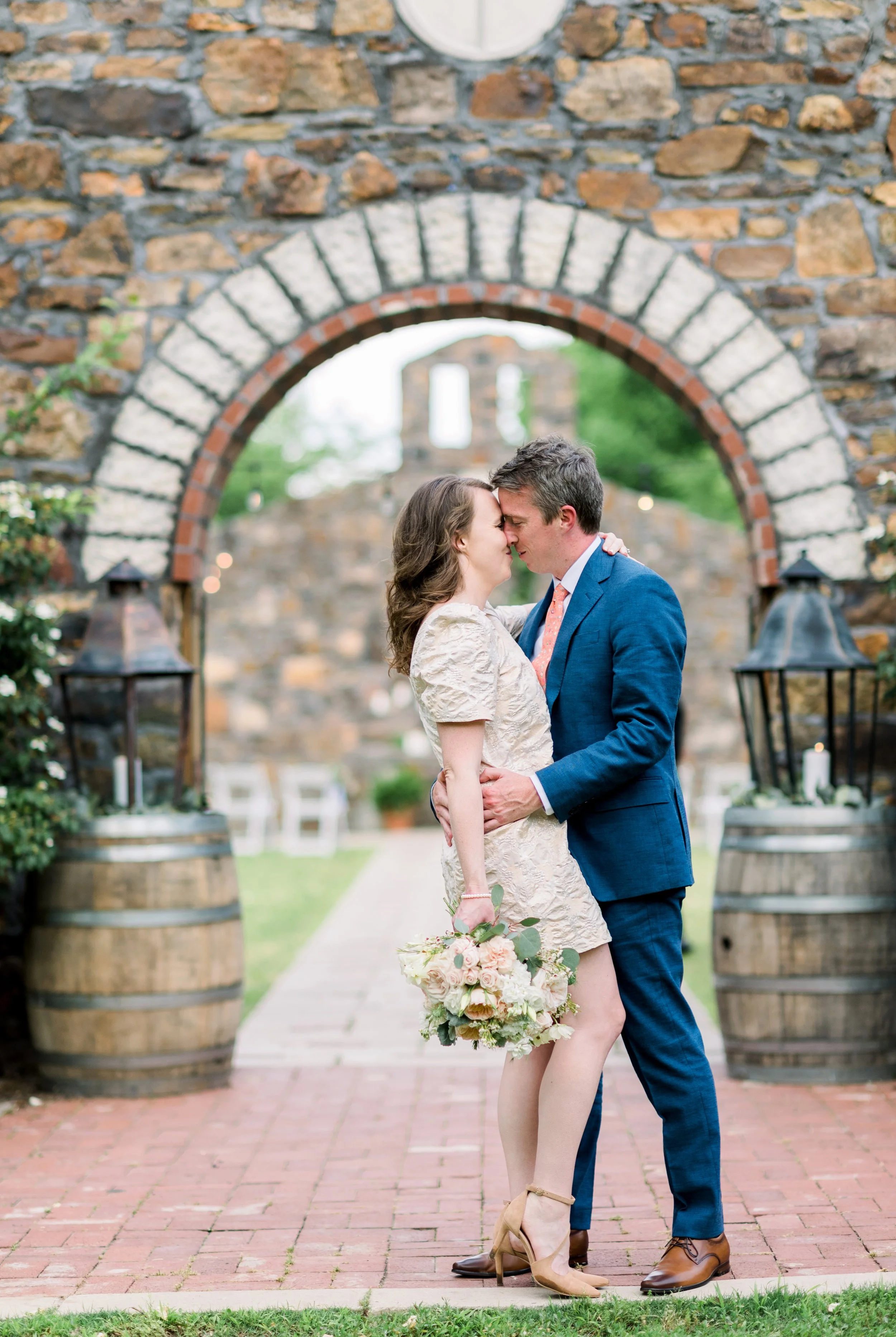 A couple embraces in front of a stone archway, with a man in a blue suit and a woman in a beige dress holding a bouquet, during a wedding celebration outdoors.