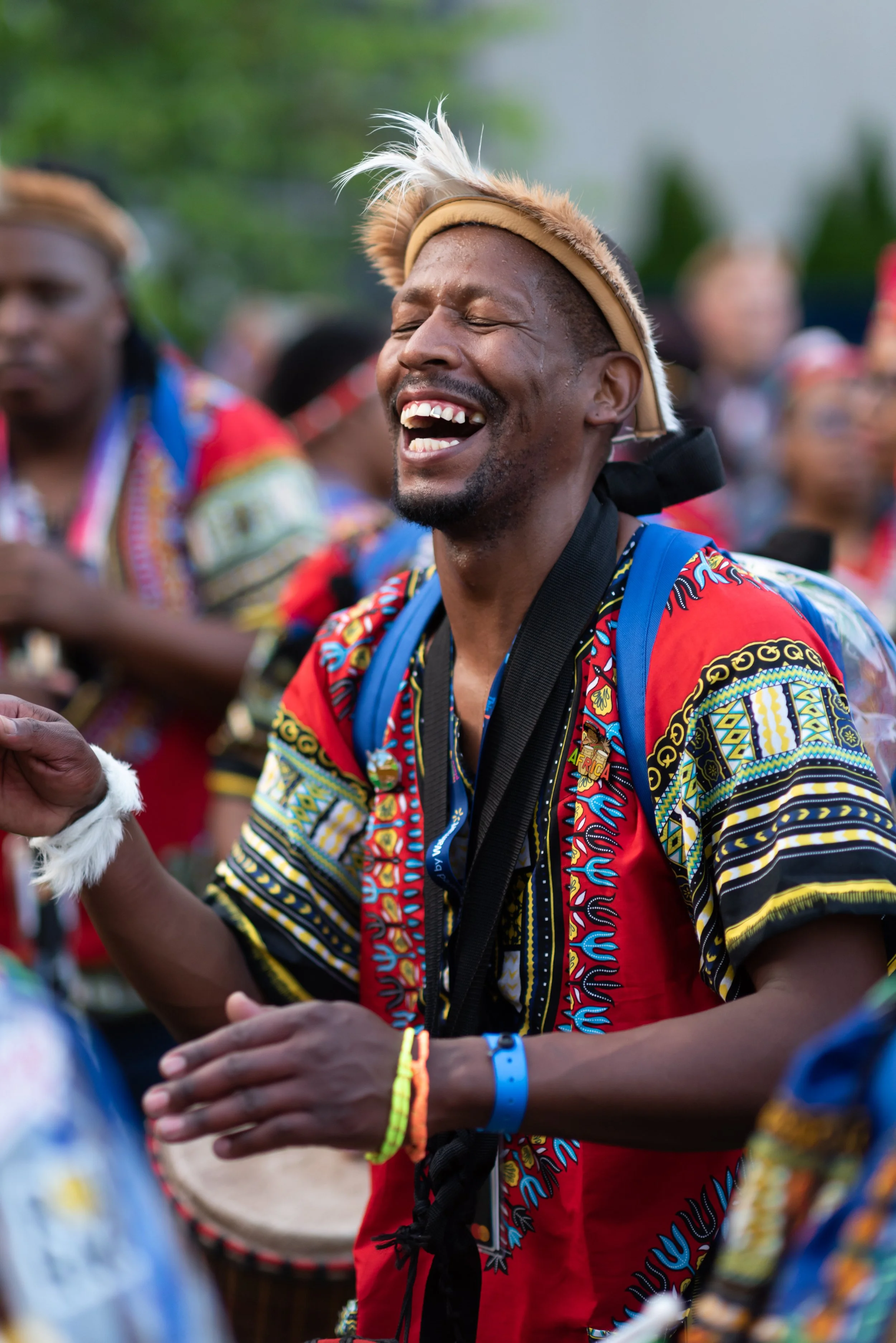 A smiling man wearing a colorful traditional African shirt and a headband, playing drums during a cultural celebration or event.