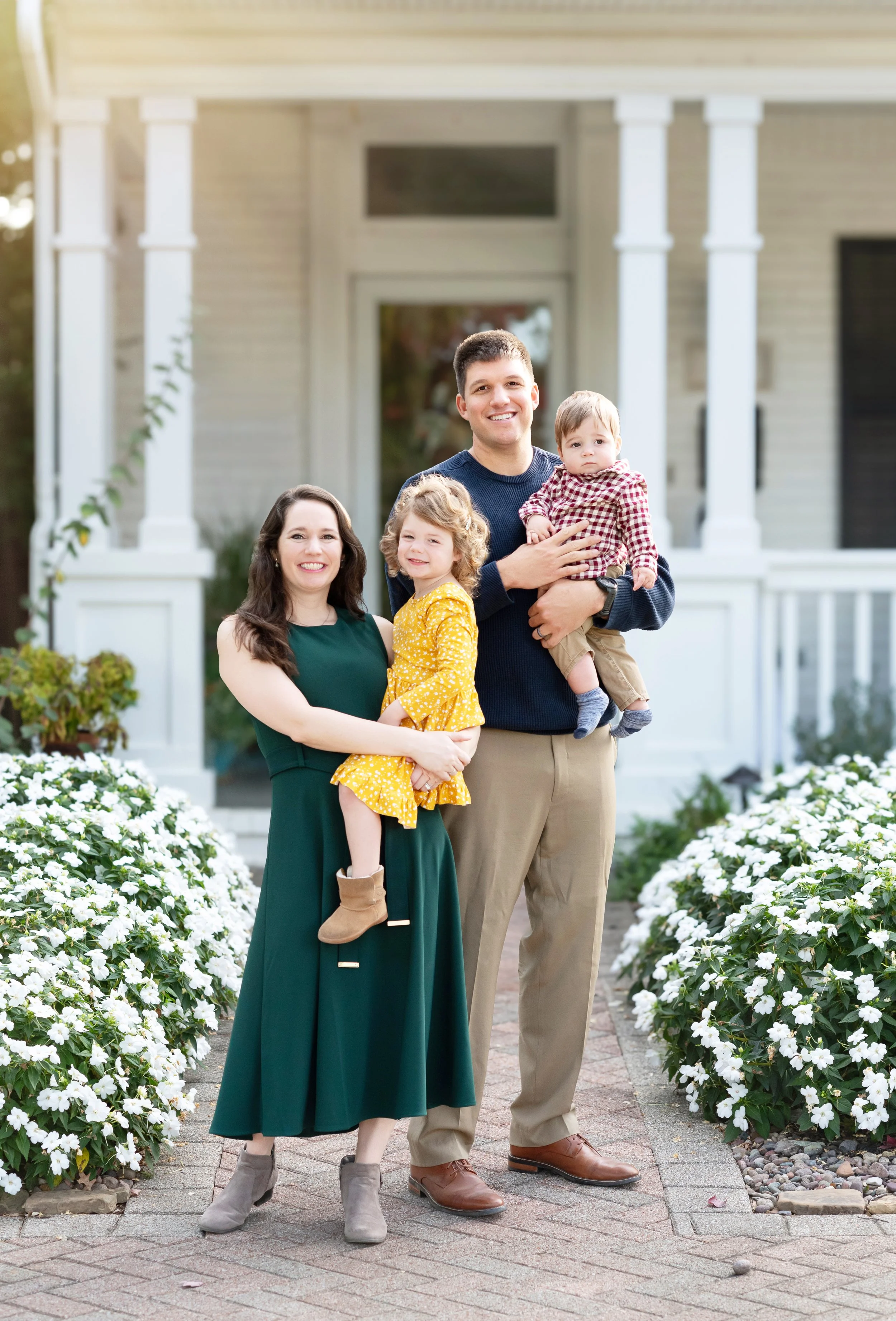 A family of four standing on a brick pathway in front of a white house with white flowers on either side. The mother is holding a young girl in a yellow dress, and the father is holding a young boy in a red checkered shirt. All are smiling.