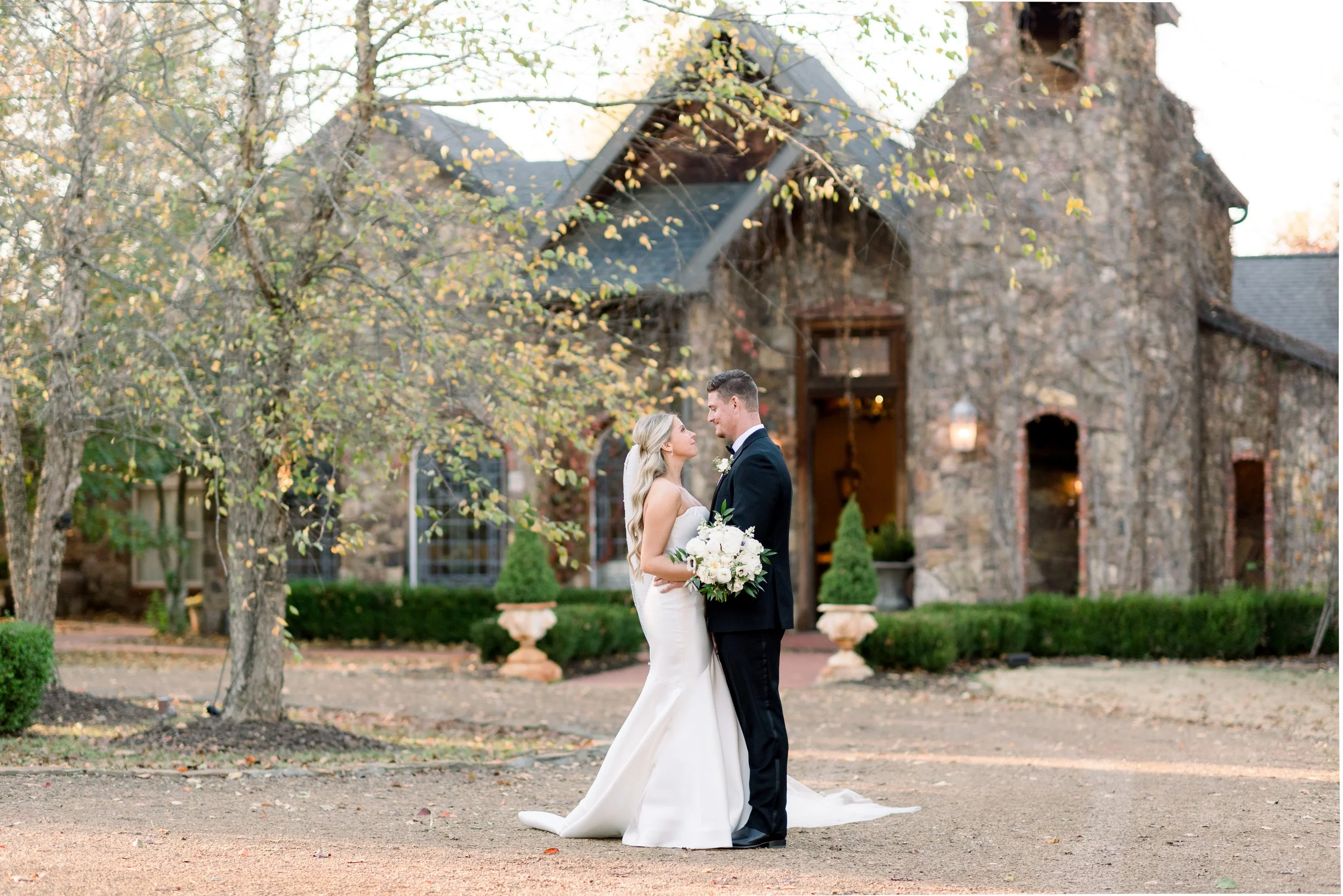 A bride and groom stand close together outside a stone building, gazing into each other's eyes on their wedding day. The bride is holding a bouquet of white flowers, wearing a white dress, and the groom is in a black tuxedo. Trees and greenery frame 