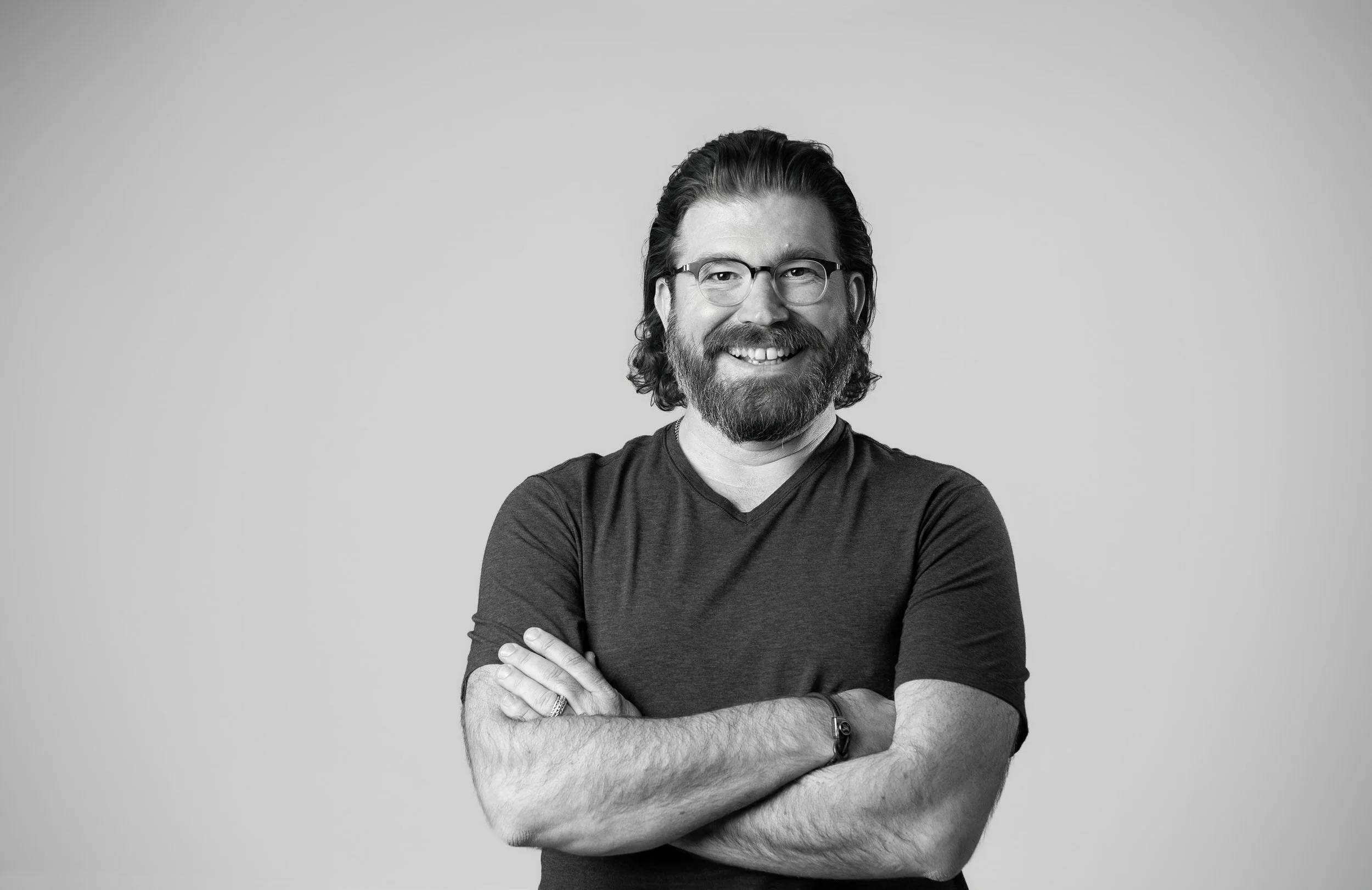 Black and white portrait of a smiling man with glasses, a beard, and long hair, standing with arms crossed against a plain background.