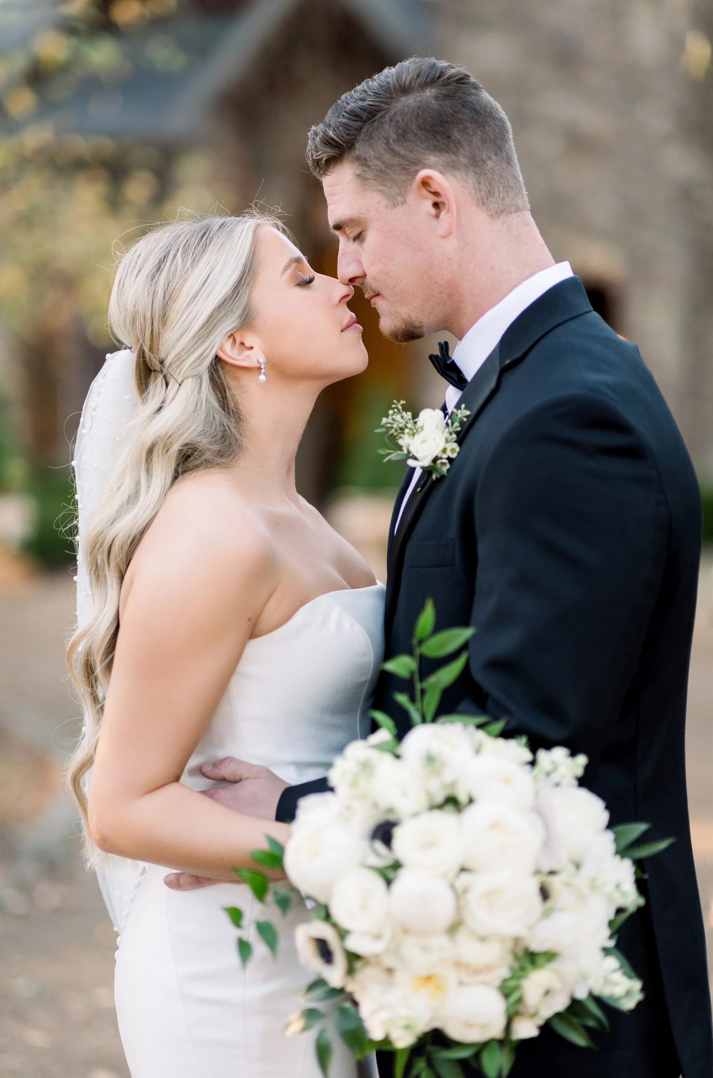 Bride and groom close together, forehead touching, outdoors wedding, bride holding bouquet of white flowers, groom in tuxedo with boutonniere.
