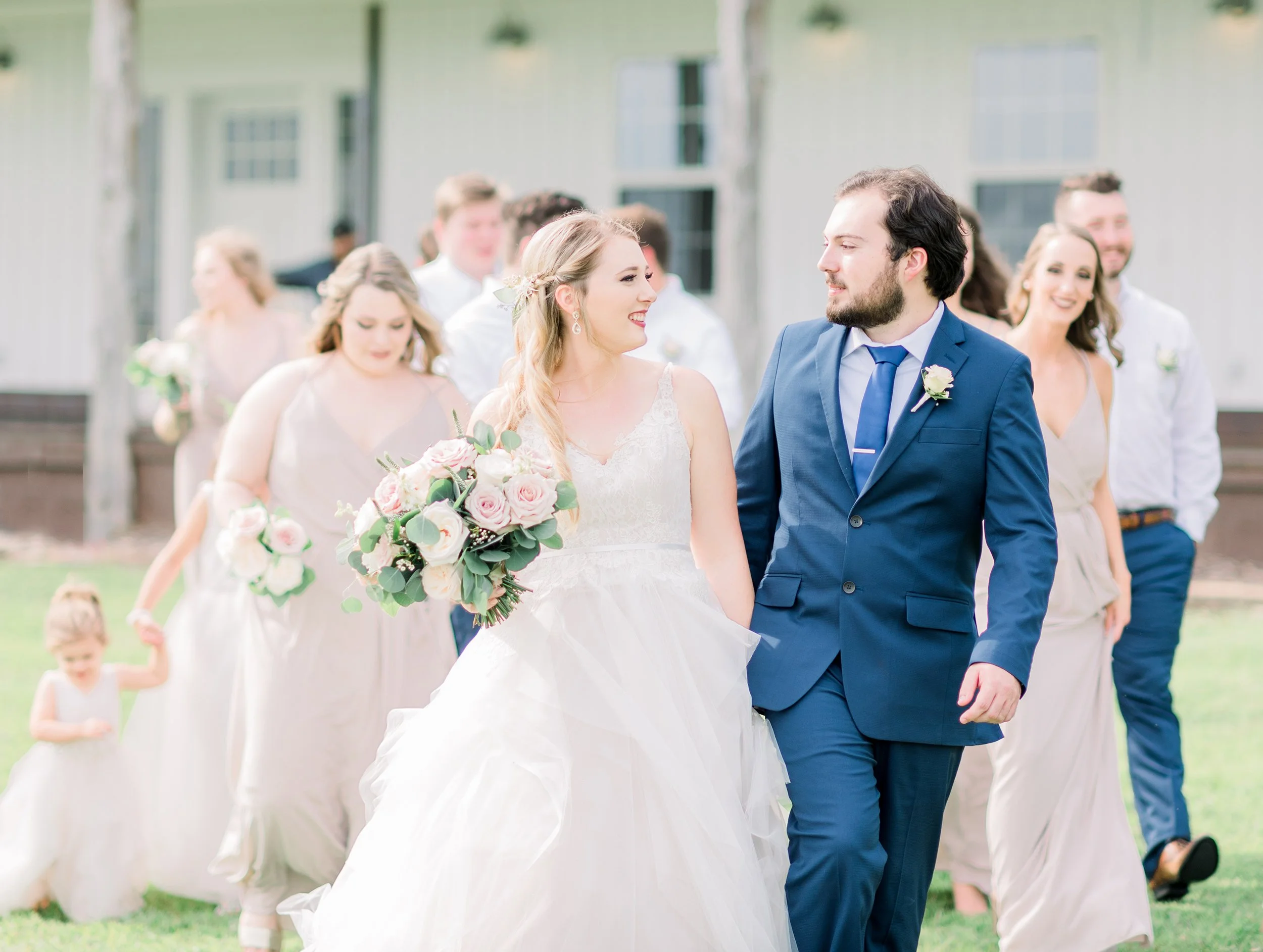 A bride and groom walking together outdoors at their wedding, surrounded by bridesmaids and groomsmen, with a bright, cheerful atmosphere.