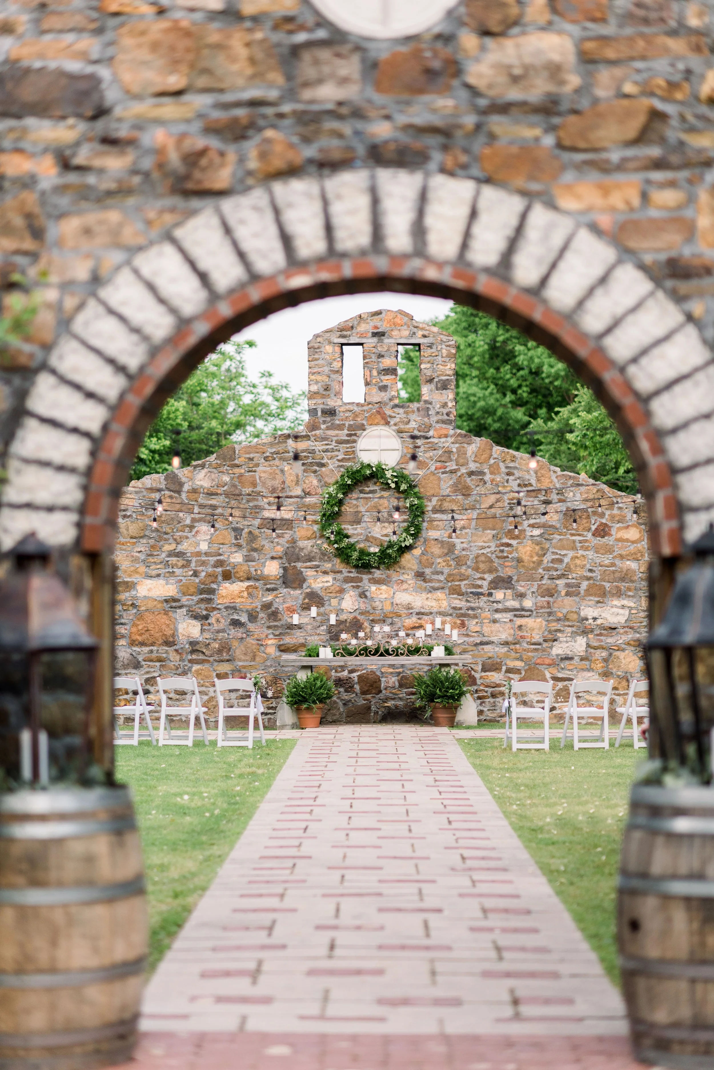 View through a brick archway into an outdoor wedding ceremony setup. The scene features a stone wall with a large green wreath, candle holders, and string lights. White folding chairs are arranged on both sides of a brick aisle, leading to the wall d