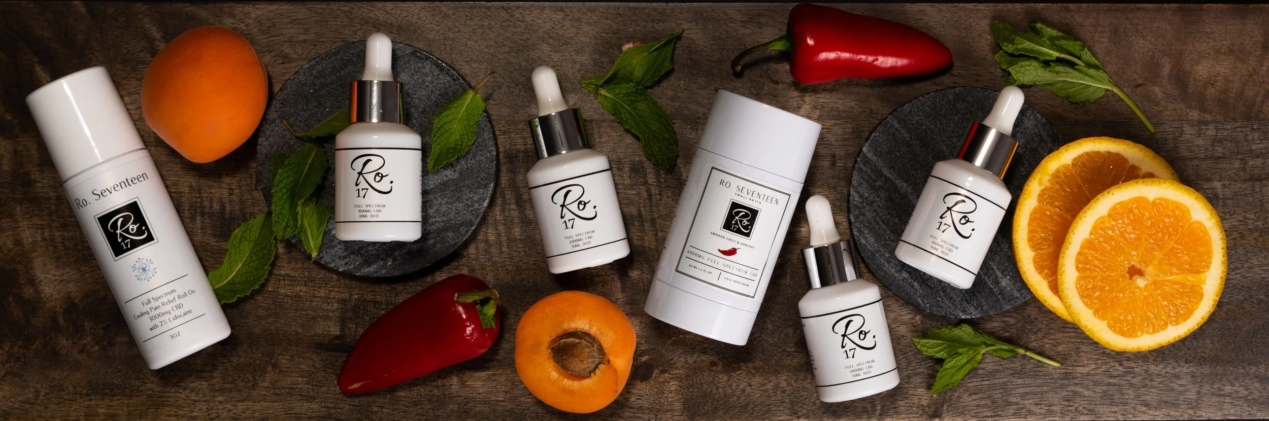 Various skincare products including bottles and droppers, fresh apricots, red and orange peppers, mint leaves, and orange slices arranged on a wooden surface.
