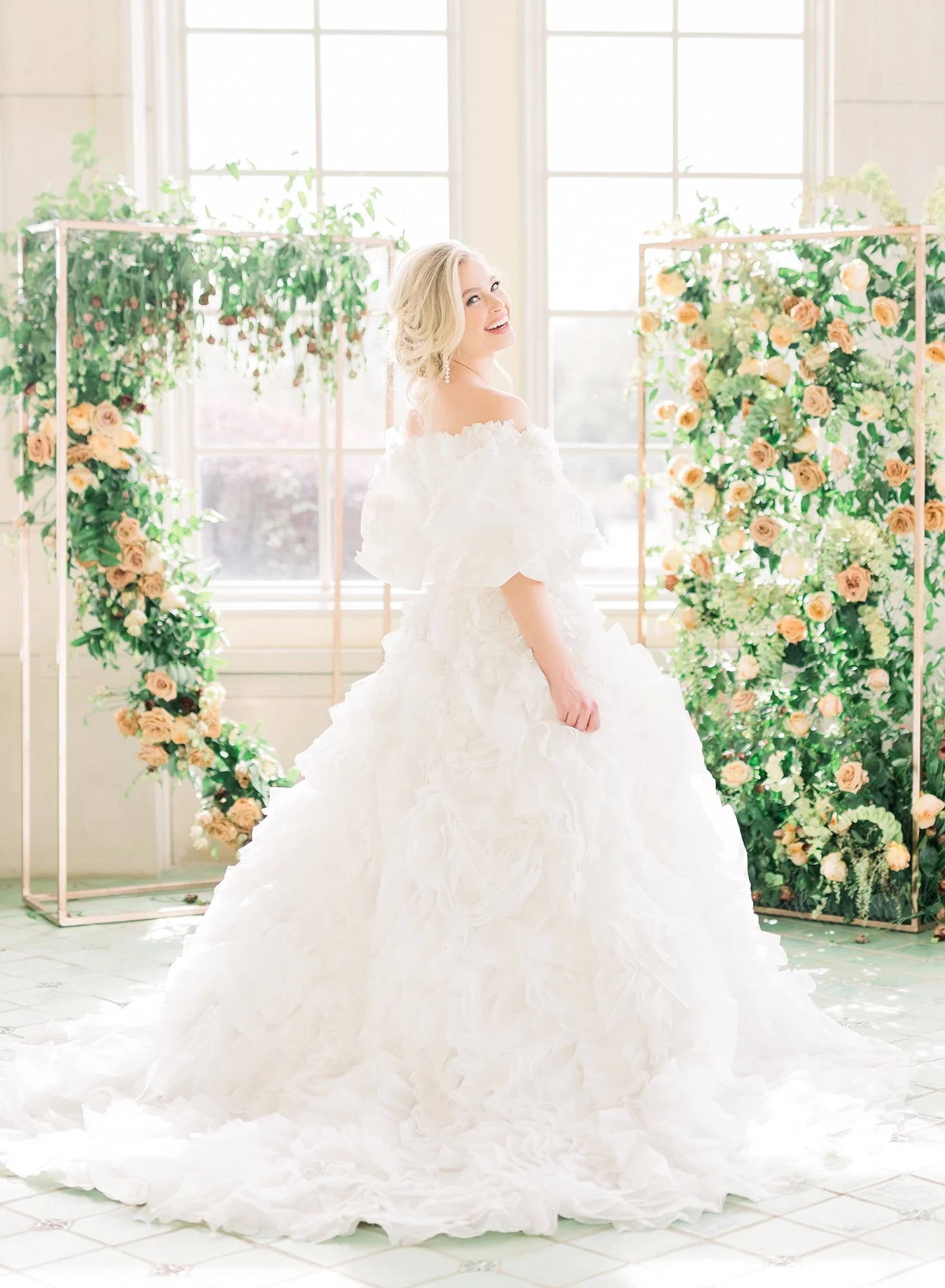 A bride in a white wedding dress with ruffles and off-the-shoulder sleeves standing indoors near large windows, smiling with a backdrop of floral arrangements of roses and greenery.