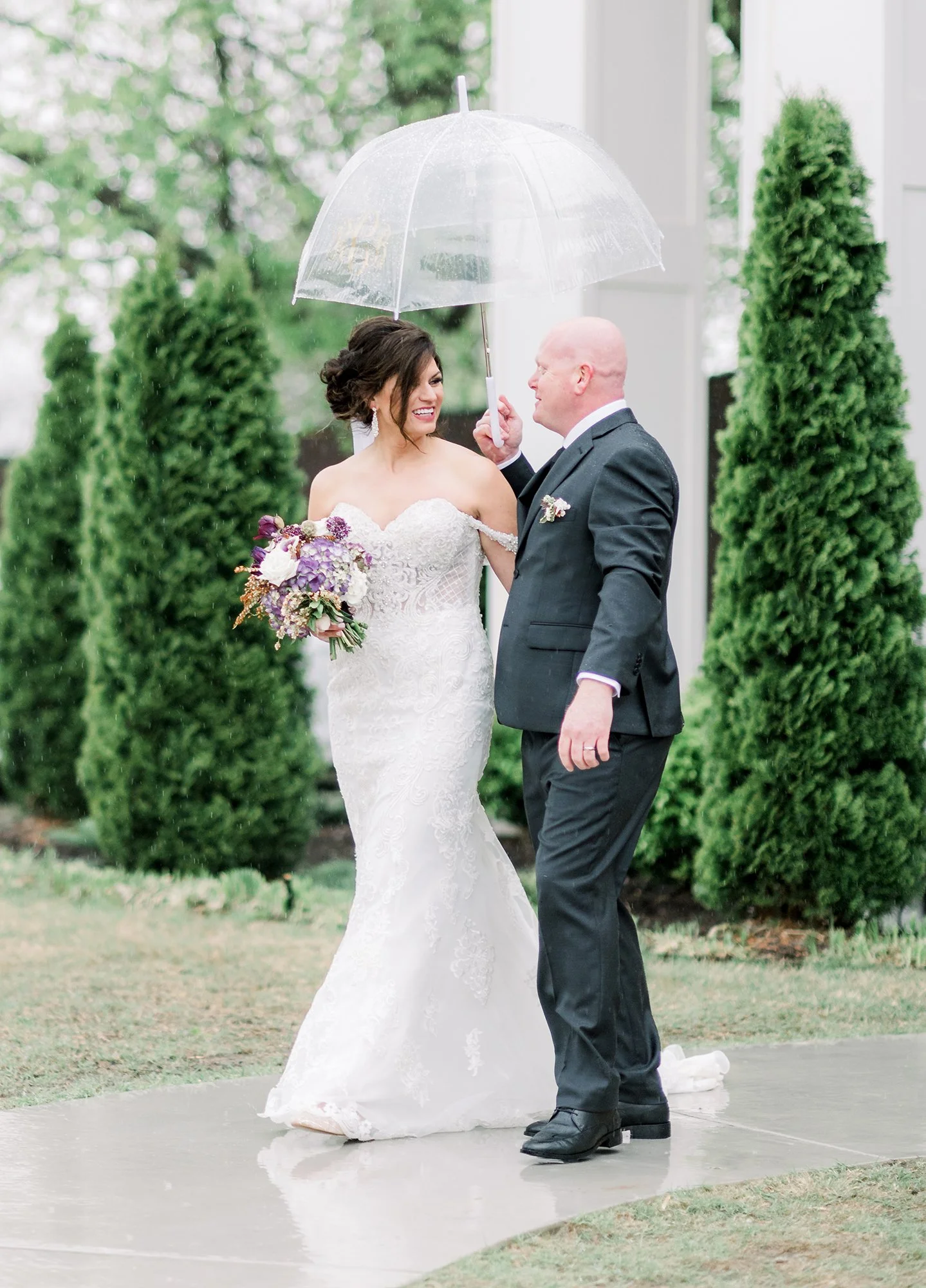 A bride and groom share a moment under a transparent umbrella outside on a rainy day. The bride is in a lace wedding gown holding a bouquet, and the groom is in a dark suit.