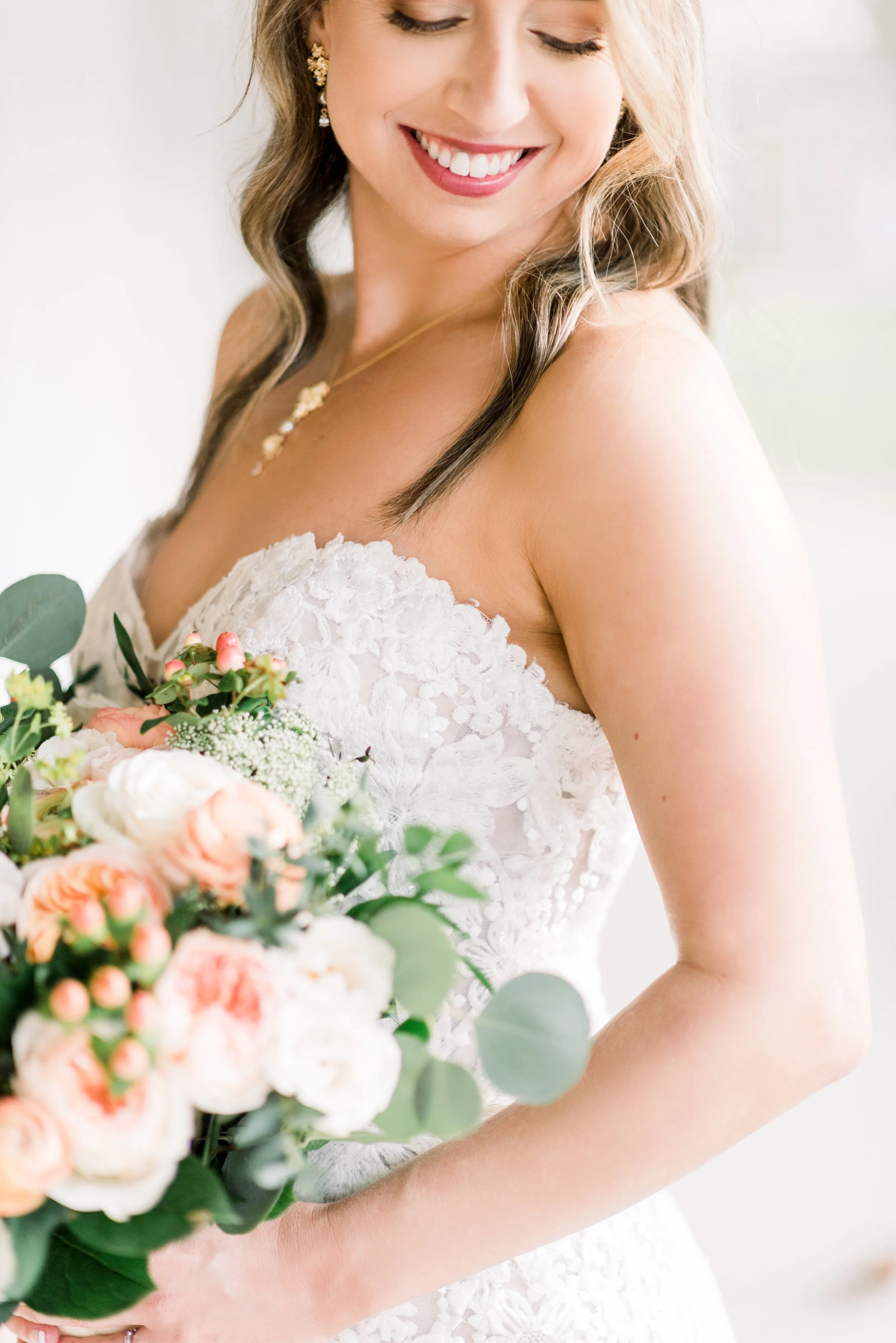 A bride in a white lace wedding dress holding a bouquet of light pink and white flowers, smiling with her eyes closed.
