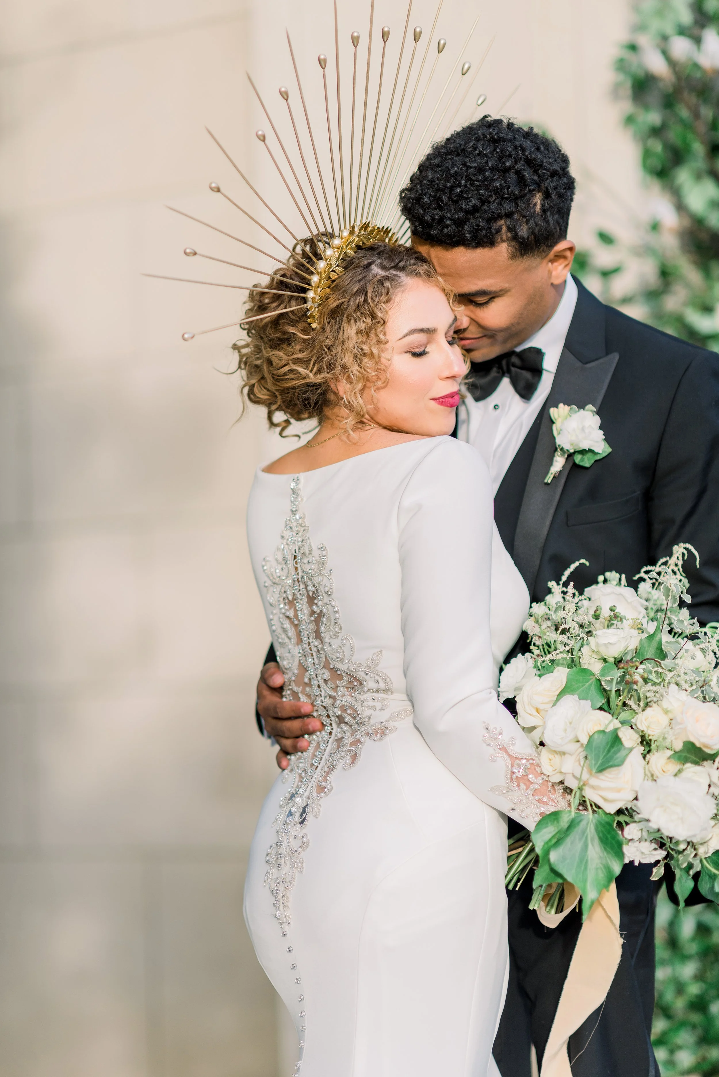 A bride and groom embracing at their wedding, with the bride wearing a white dress with intricate beading and a gold headpiece, and holding a bouquet of white flowers. The groom is dressed in a black tuxedo with a bow tie and boutonniere.