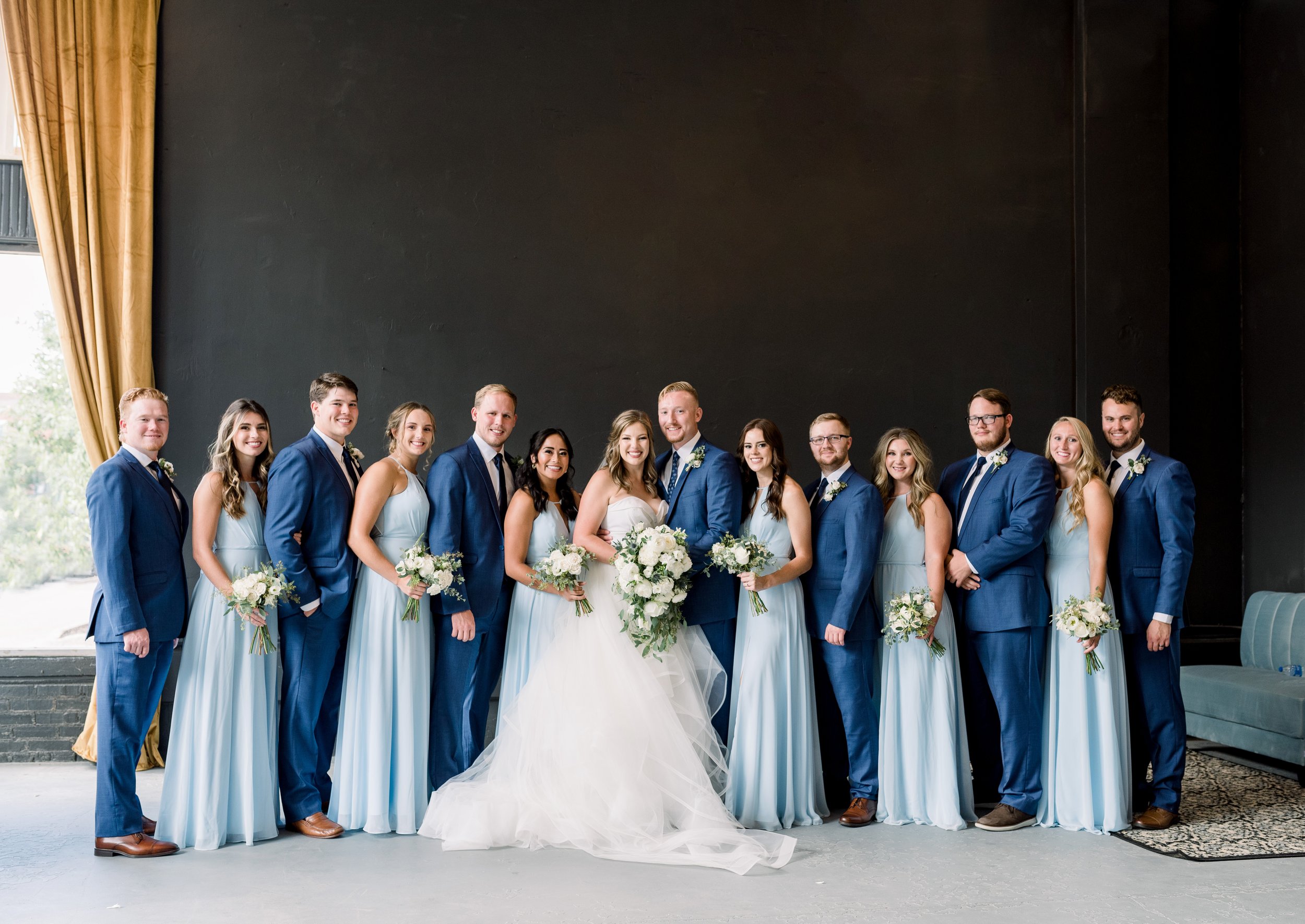 A group of 14 people, including a bride and groom in the center, posing for a wedding photo indoors. The bride is wearing a white wedding gown and holding a bouquet of flowers, while the groom is dressed in a blue suit. The bridesmaids are wearing li