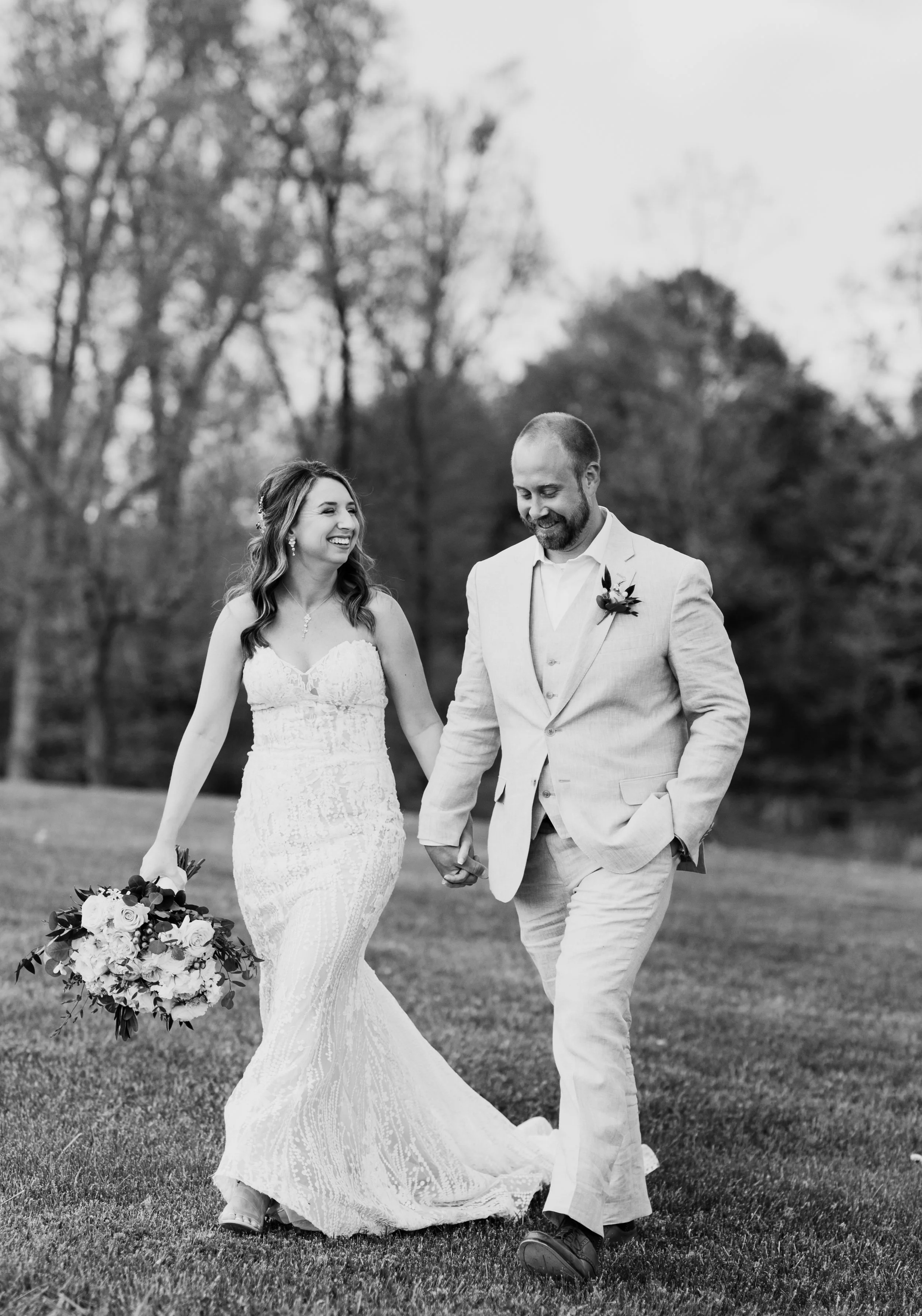 A black-and-white photo of a happy bride and groom walking hand in hand outdoors, smiling, with trees in the background.