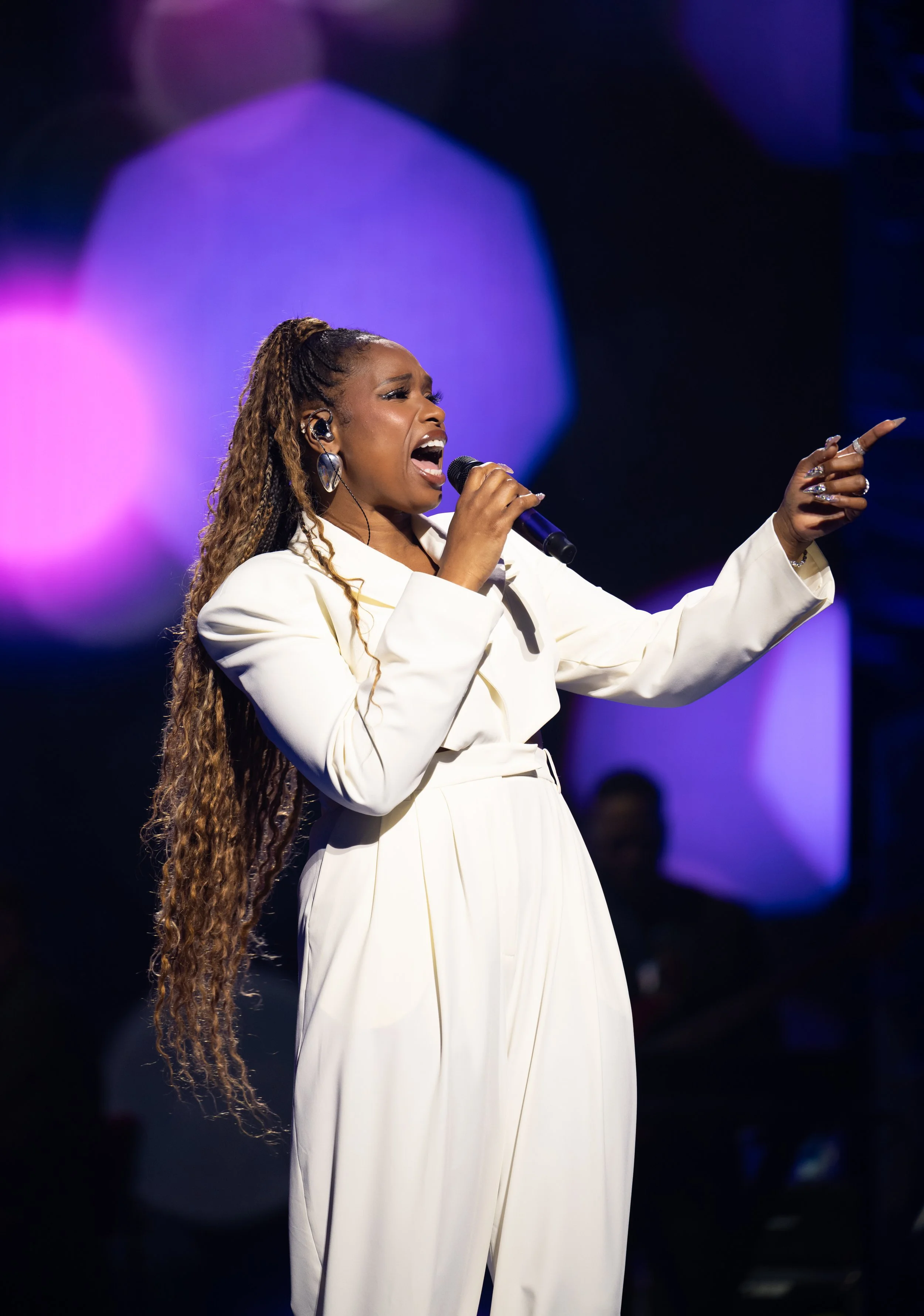 A woman with long, curly hair singing into a microphone on stage, wearing a white outfit, with colorful blurred lights in the background.