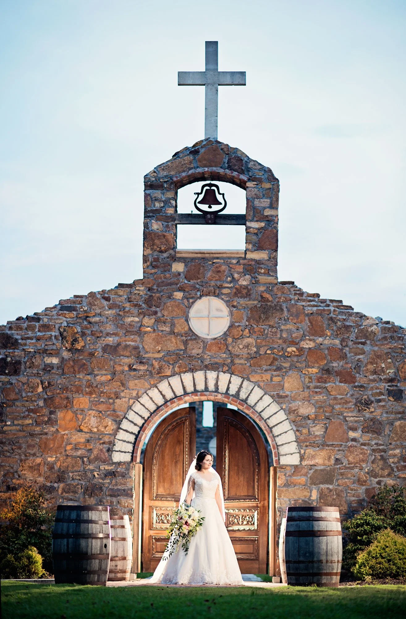 A bride in a white wedding dress holding a bouquet stands in front of a stone church with wooden doors and a bell tower topped with a cross.