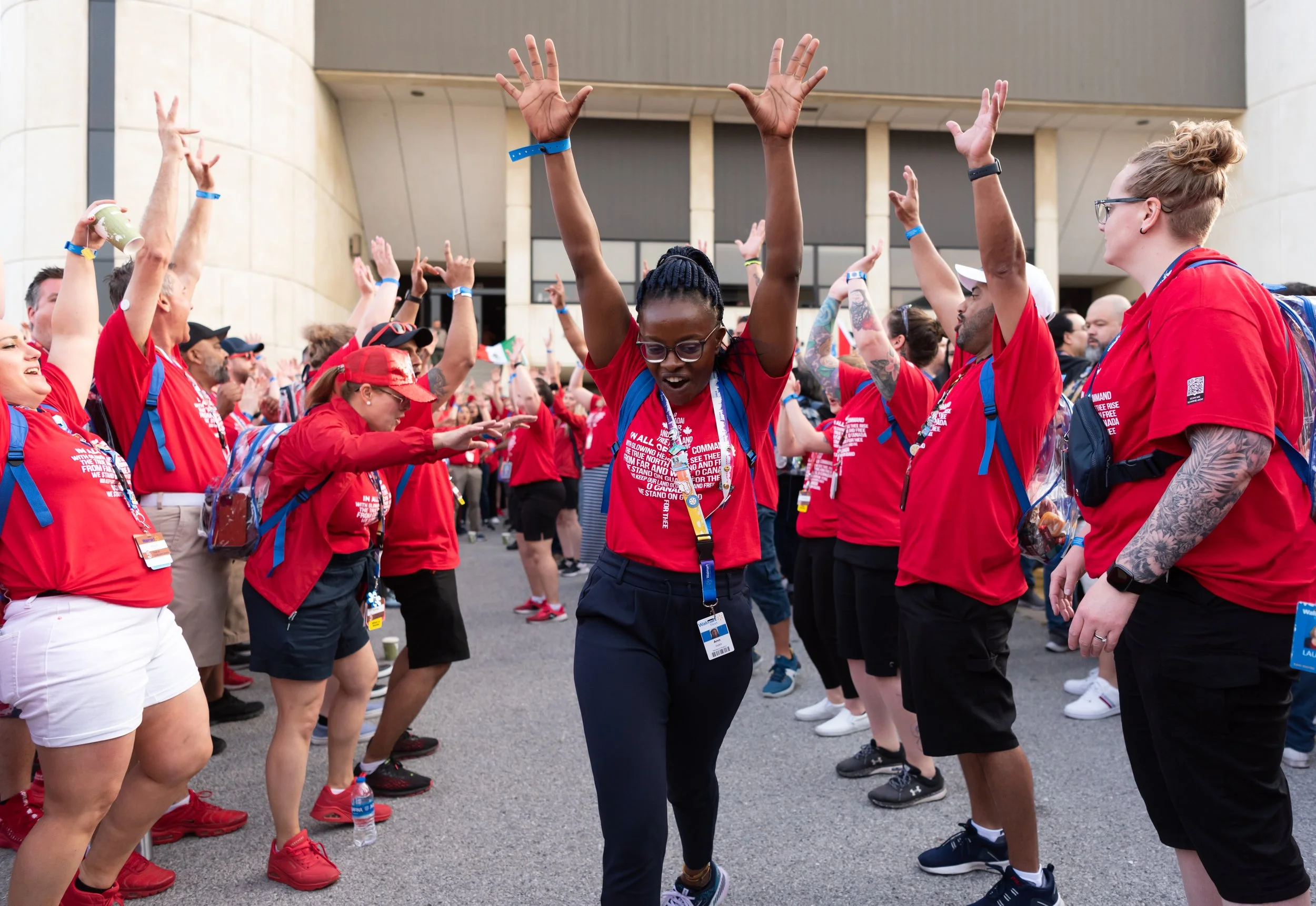 Group of people wearing red shirts and blue lanyards, participating in a celebratory outdoor event, some with arms raised, one person dancing in the center.