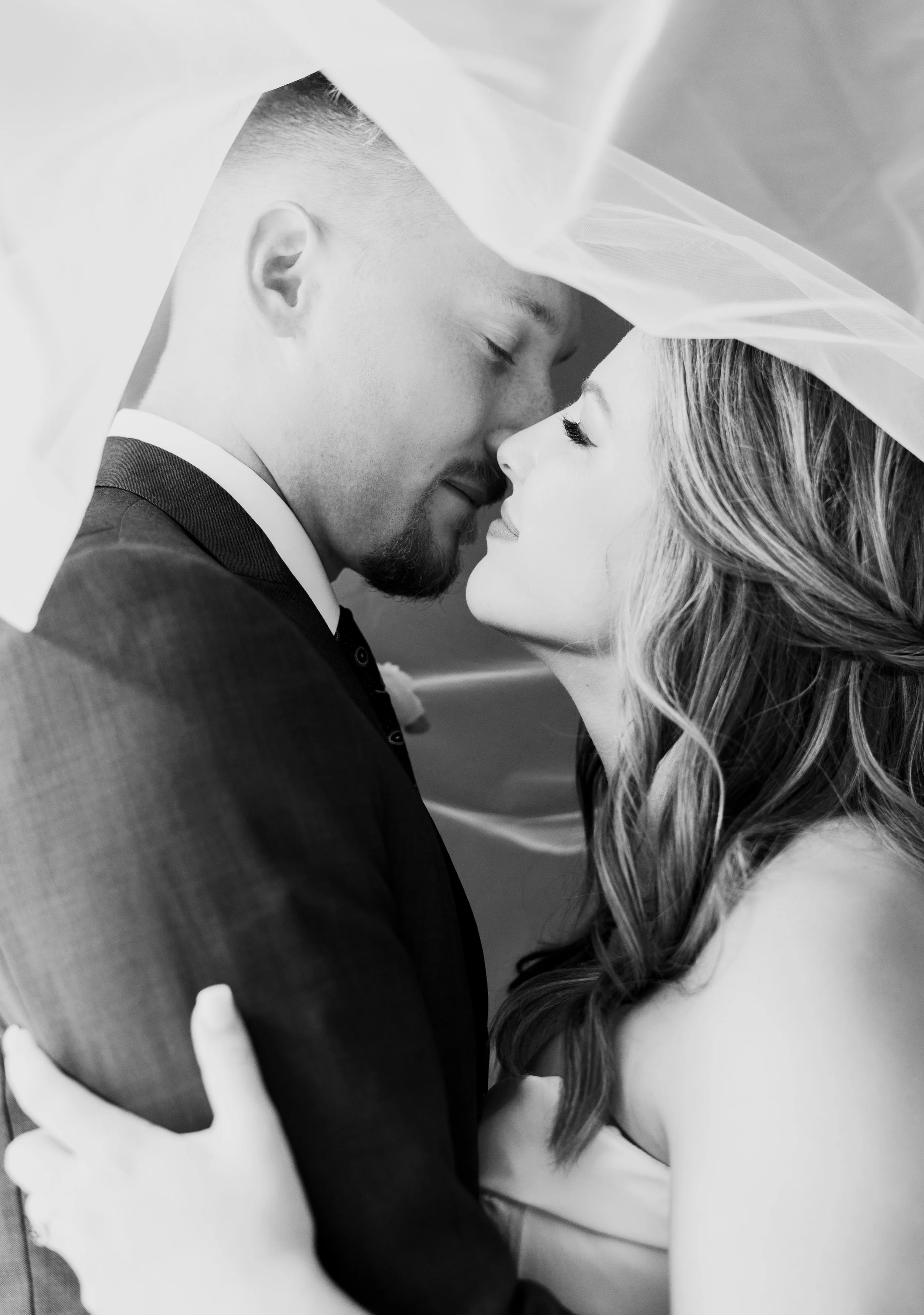 A black and white photo of a bride and groom close together, with their foreheads and noses touching, under a veil.