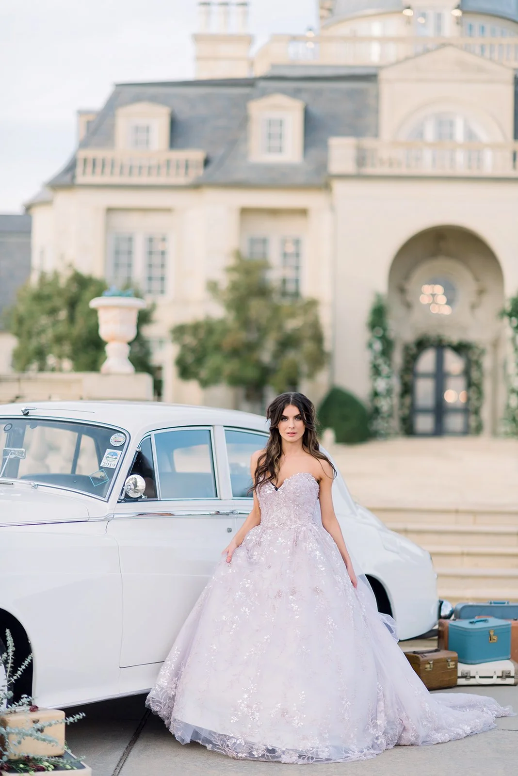 A young woman in a strapless, light purple, sequined ball gown standing next to a white vintage car outside a large, ornate house with steps and columns, with luggage and suitcases nearby.
