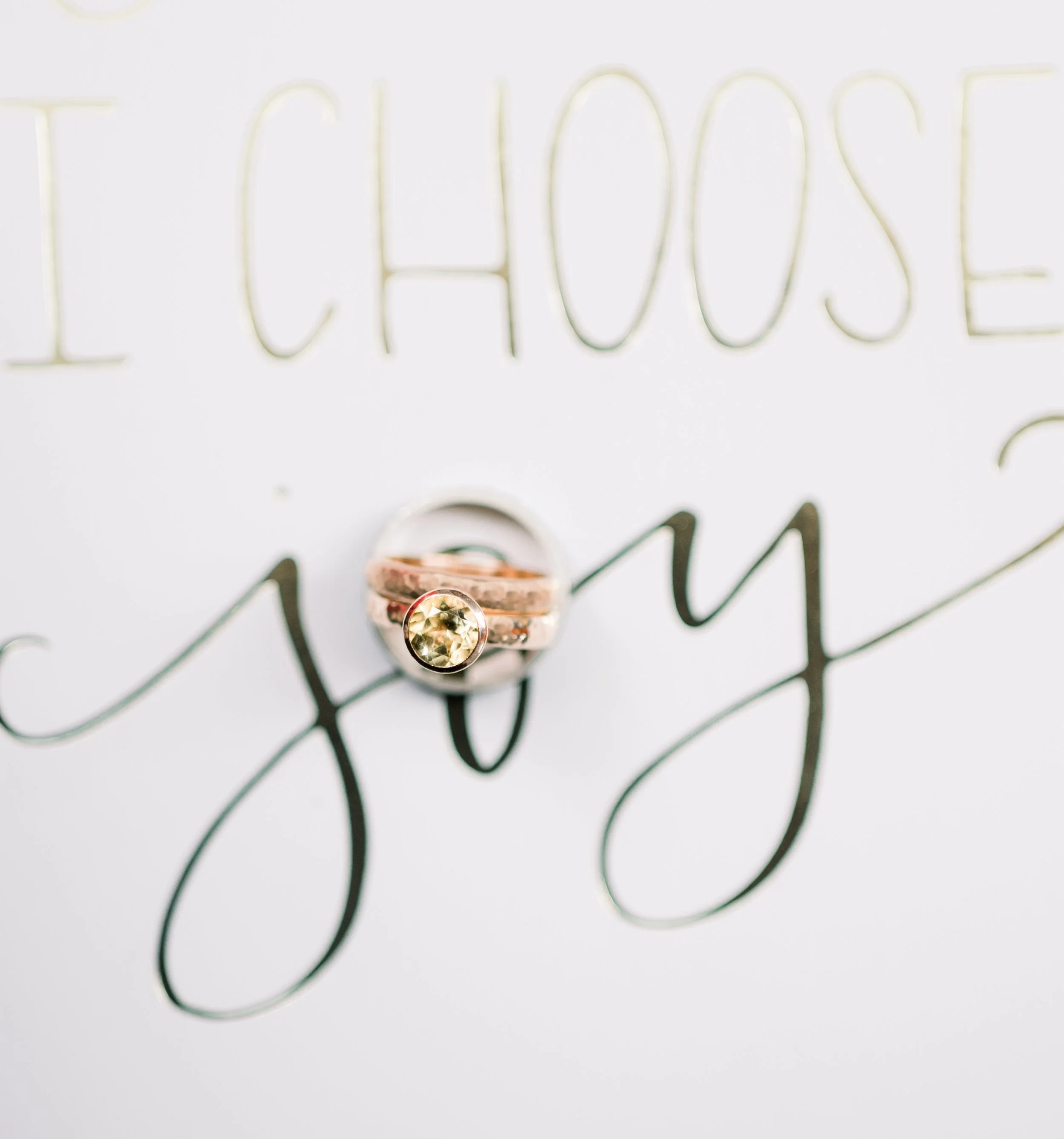 Close-up of a wedding ring with a yellow gemstone, placed on a white paper with the words 'I CHOOSE you' written in elegant black and gold script.