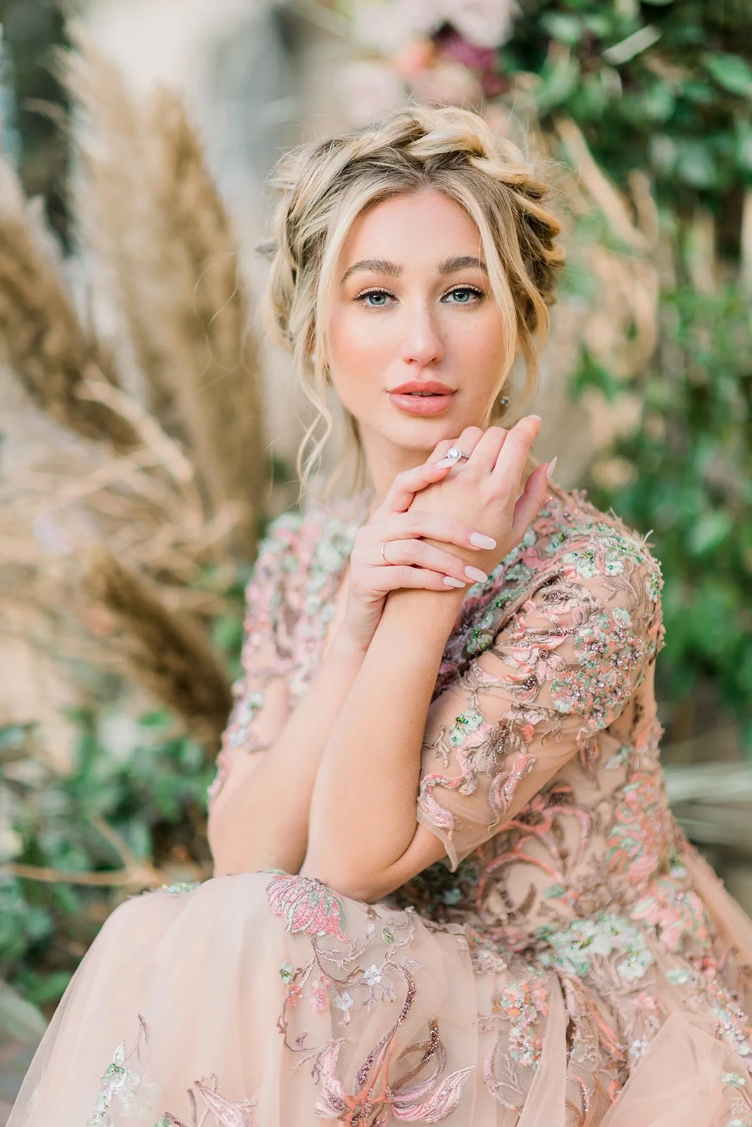 A woman with blonde hair styled in loose waves sits outdoors against a backdrop of greenery. She is wearing a floral, embroidered dress with pastel colors, and has a ring on her finger. She gazes softly at the camera with her hands gently resting nea