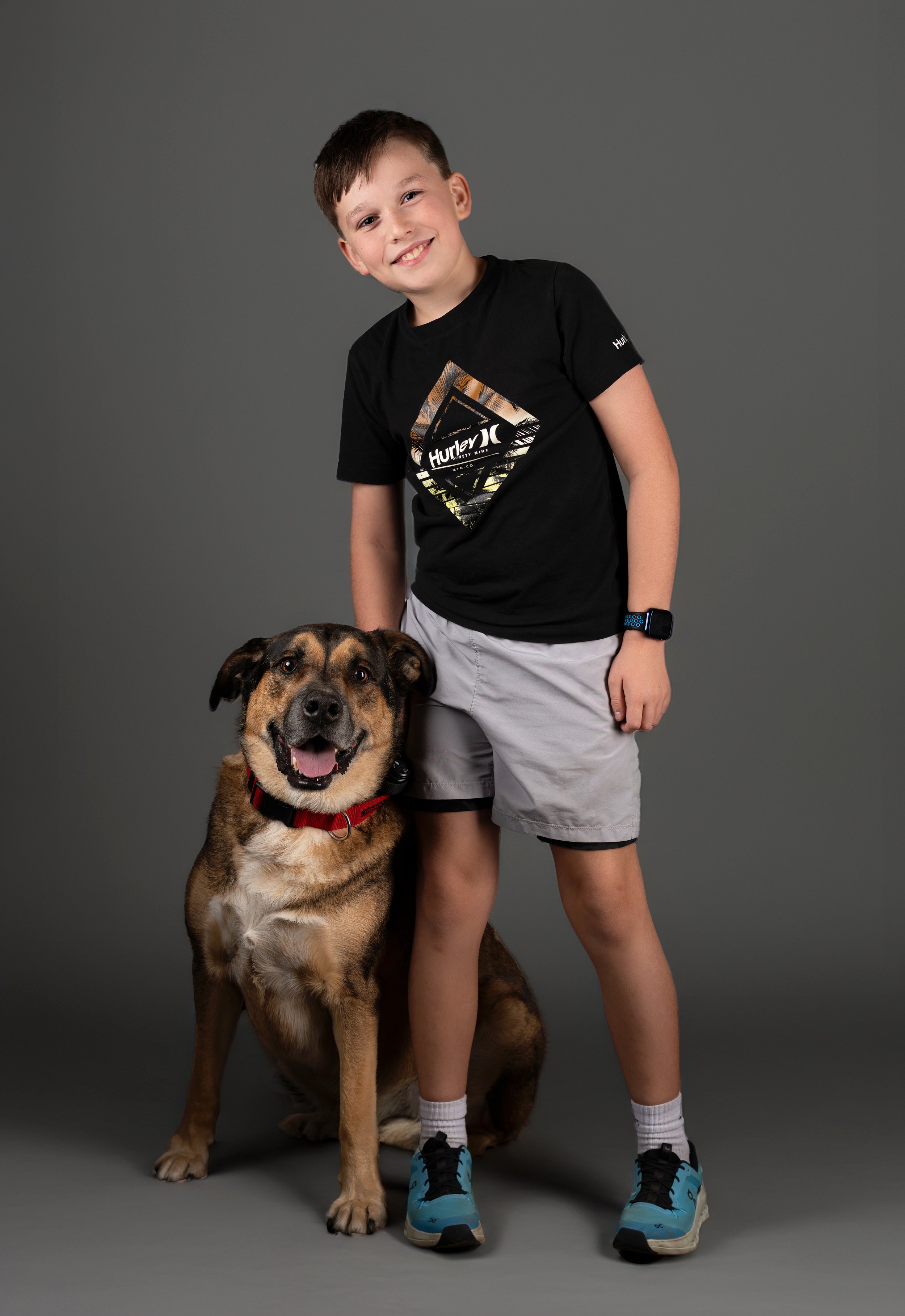 A young boy with short brown hair, smiling, wearing a black Hurley t-shirt, gray shorts, and blue sneakers, standing next to a large mixed-breed dog with brown and black fur, sitting on a gray studio backdrop.