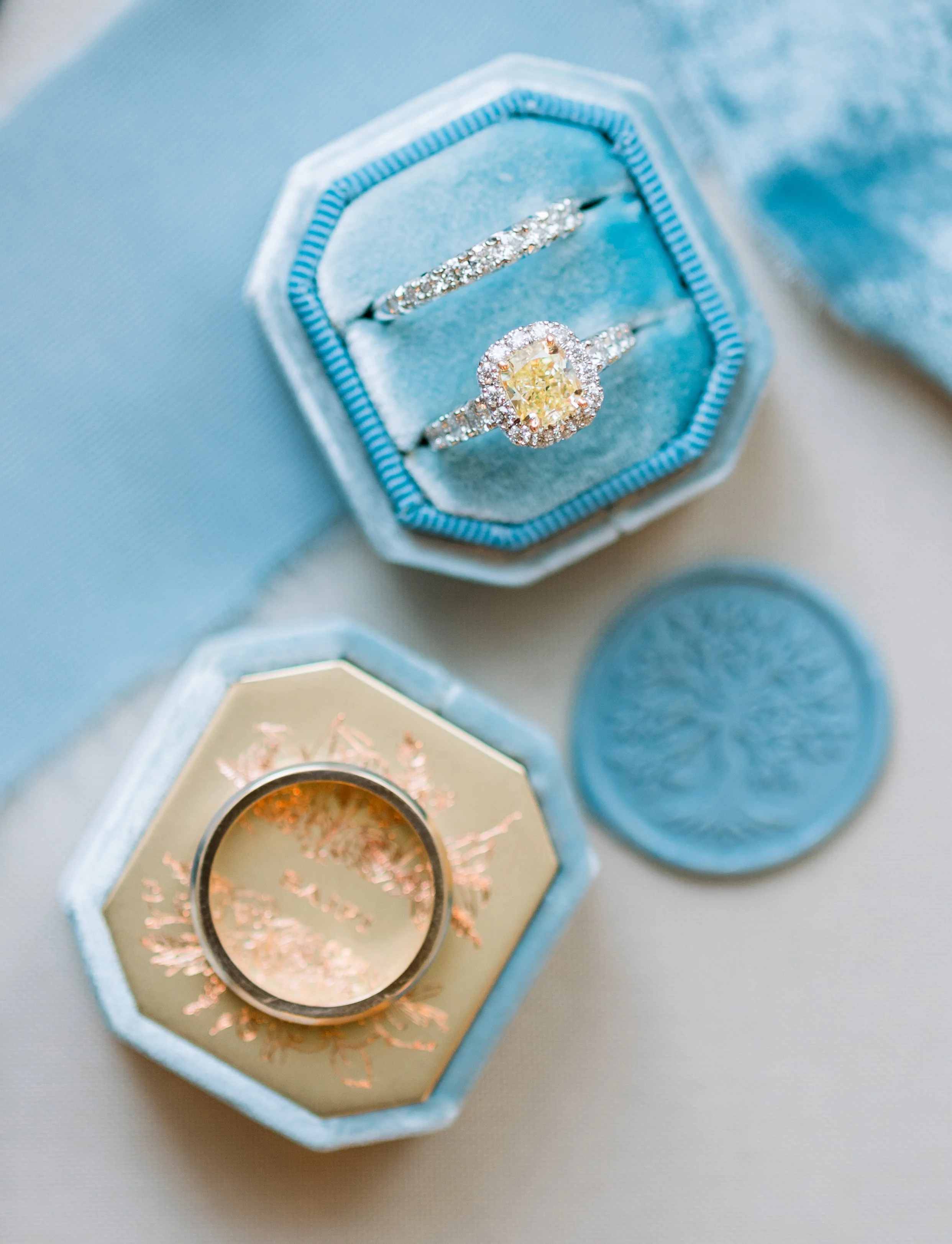 Close-up of a jewelry box containing two rings, one with a yellow gemstone surrounded by diamonds and the other with a diamond band, along with a coin and a decorative element in the background.