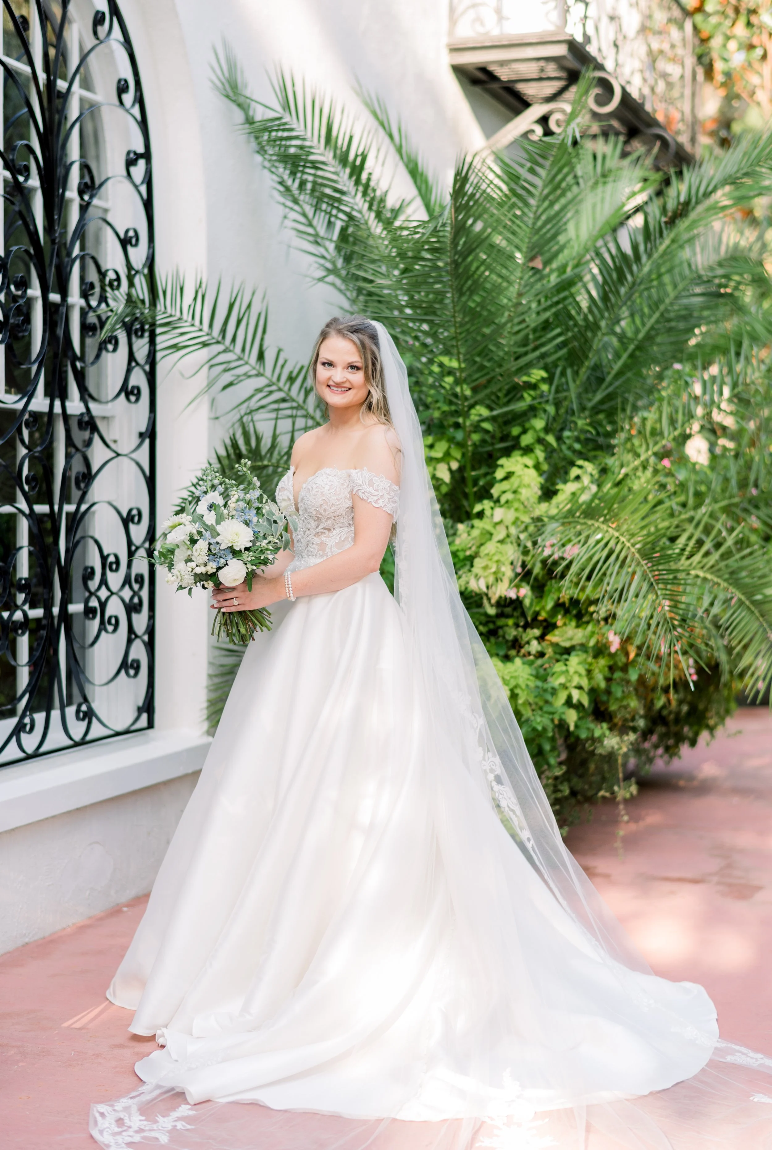 A bride in a white wedding gown holding a bouquet, standing outdoors with green tropical plants and a black decorative iron window behind her.