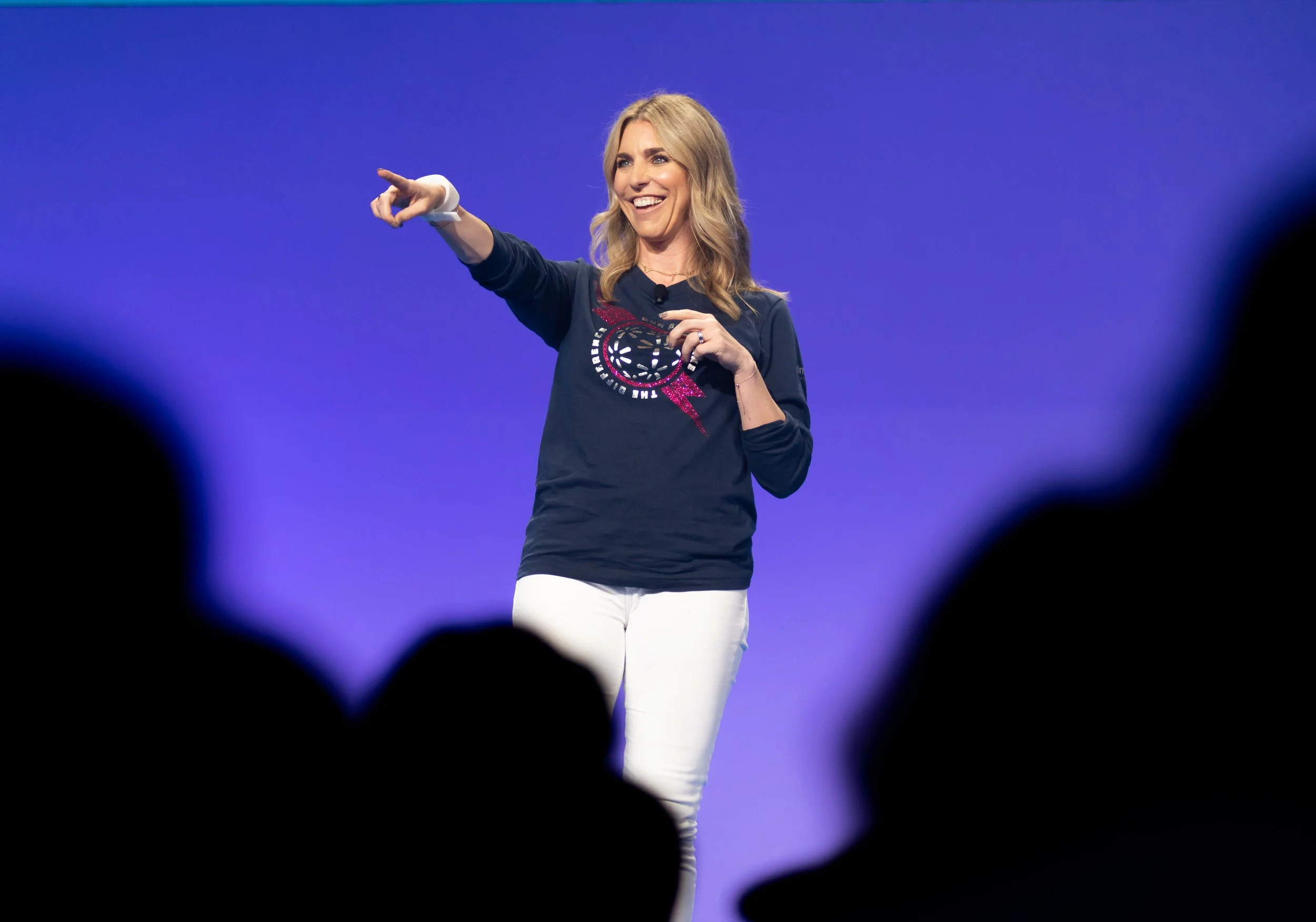 A woman with blonde hair, smiling, pointing with her right hand while standing on stage in front of a purple background, wearing a navy blue sweatshirt with graphic design and white pants, audience silhouettes in foreground.