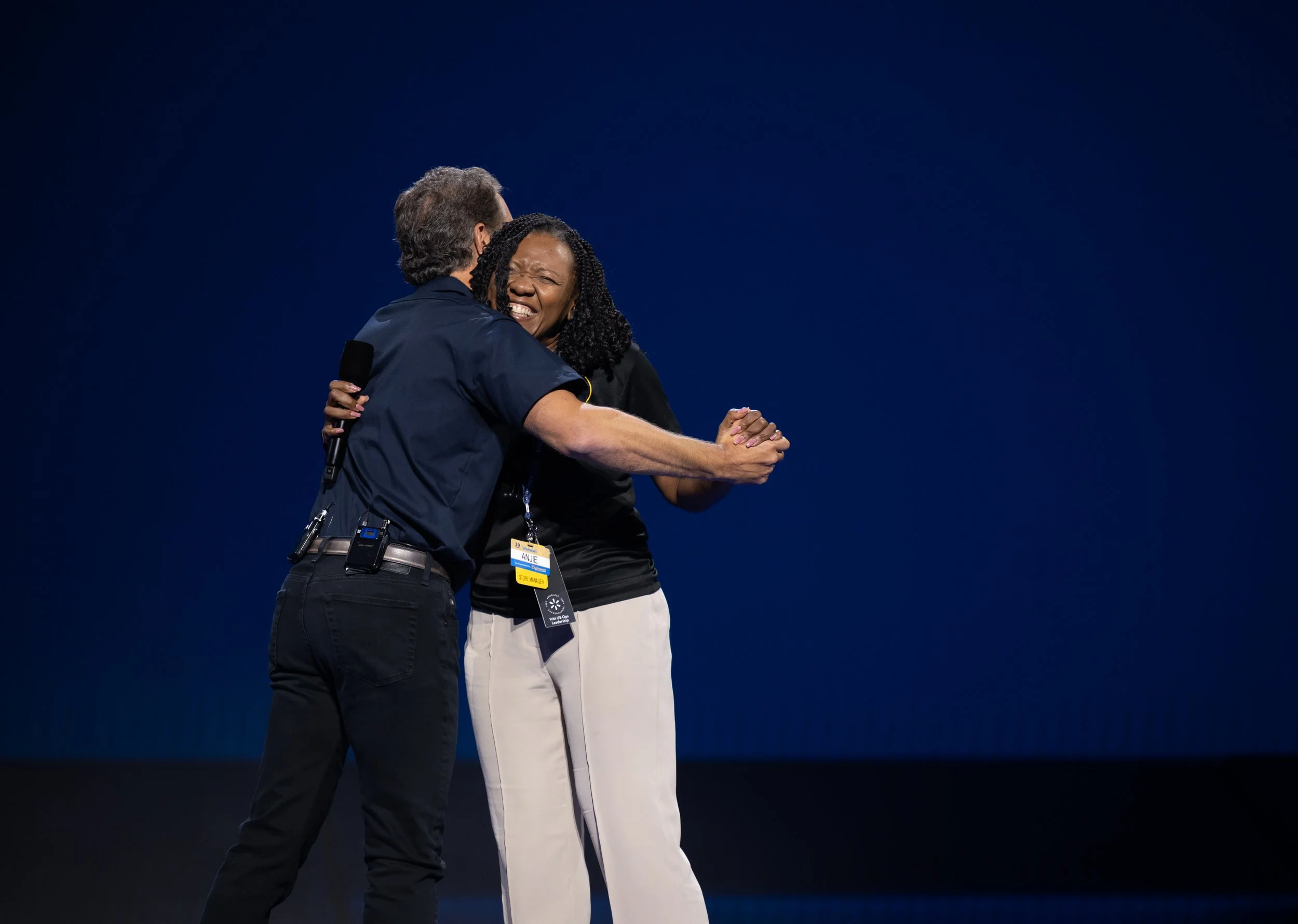 Two people hugging on stage, one man and one woman, with a dark blue background. The man is holding a microphone and wearing black pants and a black shirt, while the woman is smiling and wearing a black top and light-colored pants.