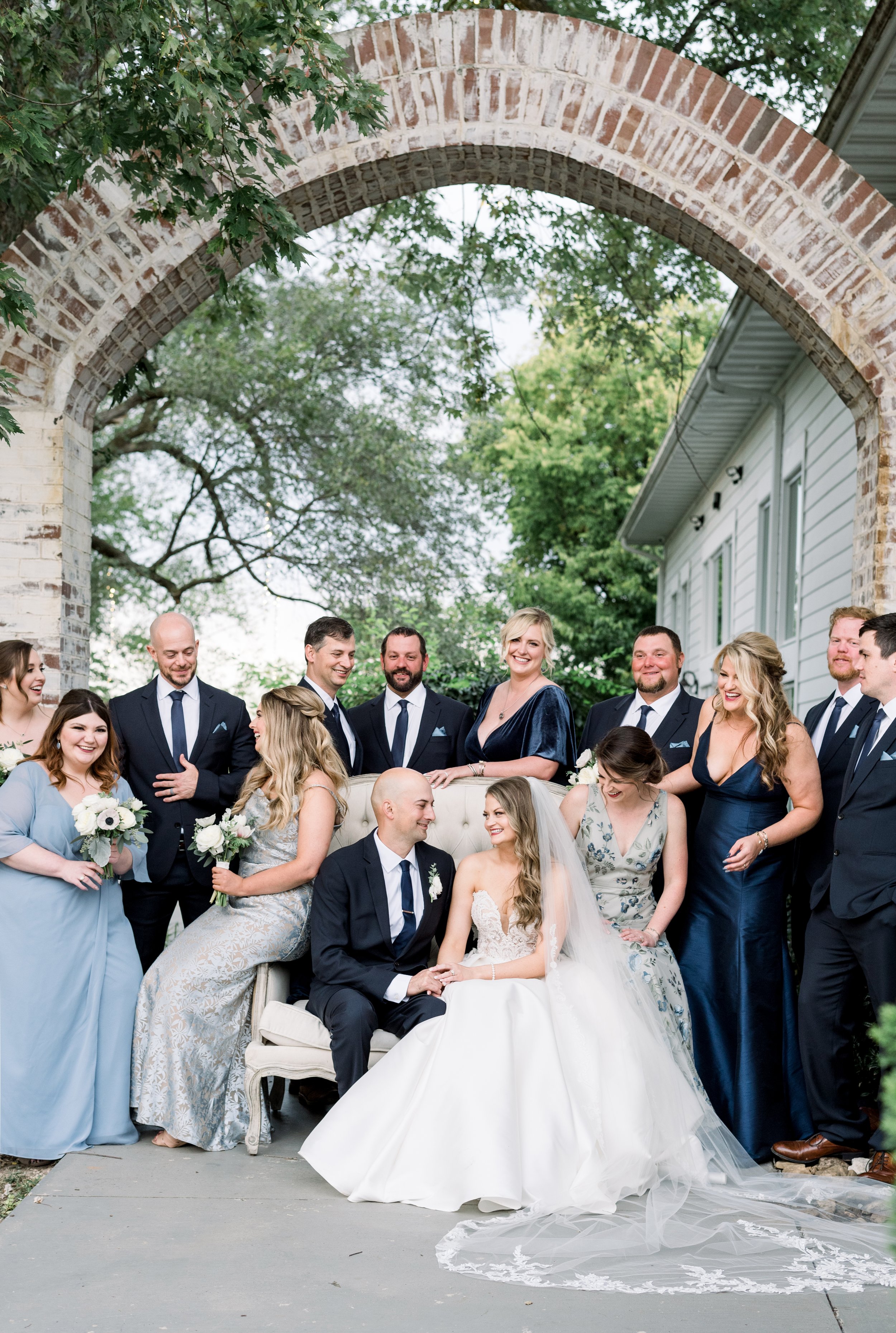 A wedding party posed under an arched brick gateway, with the bride and groom sitting on a vintage-style sofa and the bridesmaids and groomsmen standing around them, outdoors with green trees and a white house in the background.