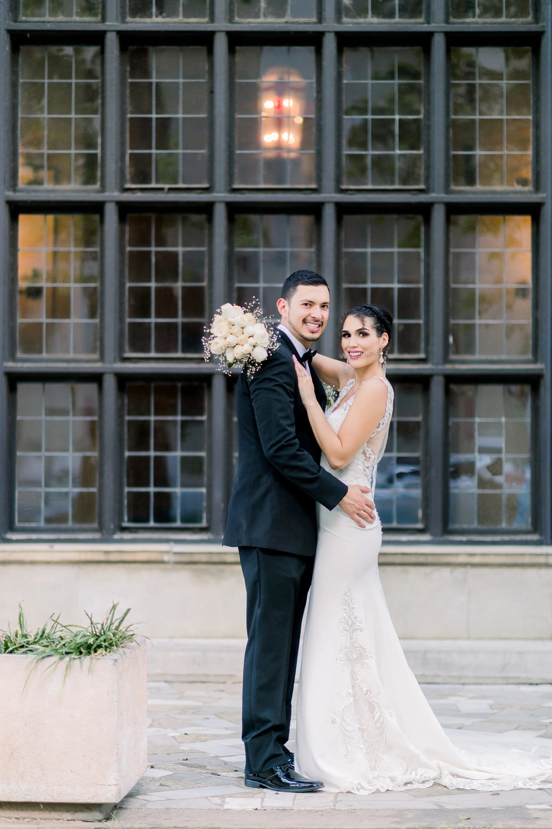 A newlywed couple in wedding attire posing outdoors in front of a large window with a reflection of a building. The groom is holding a bouquet of white flowers, and both are smiling and embracing.