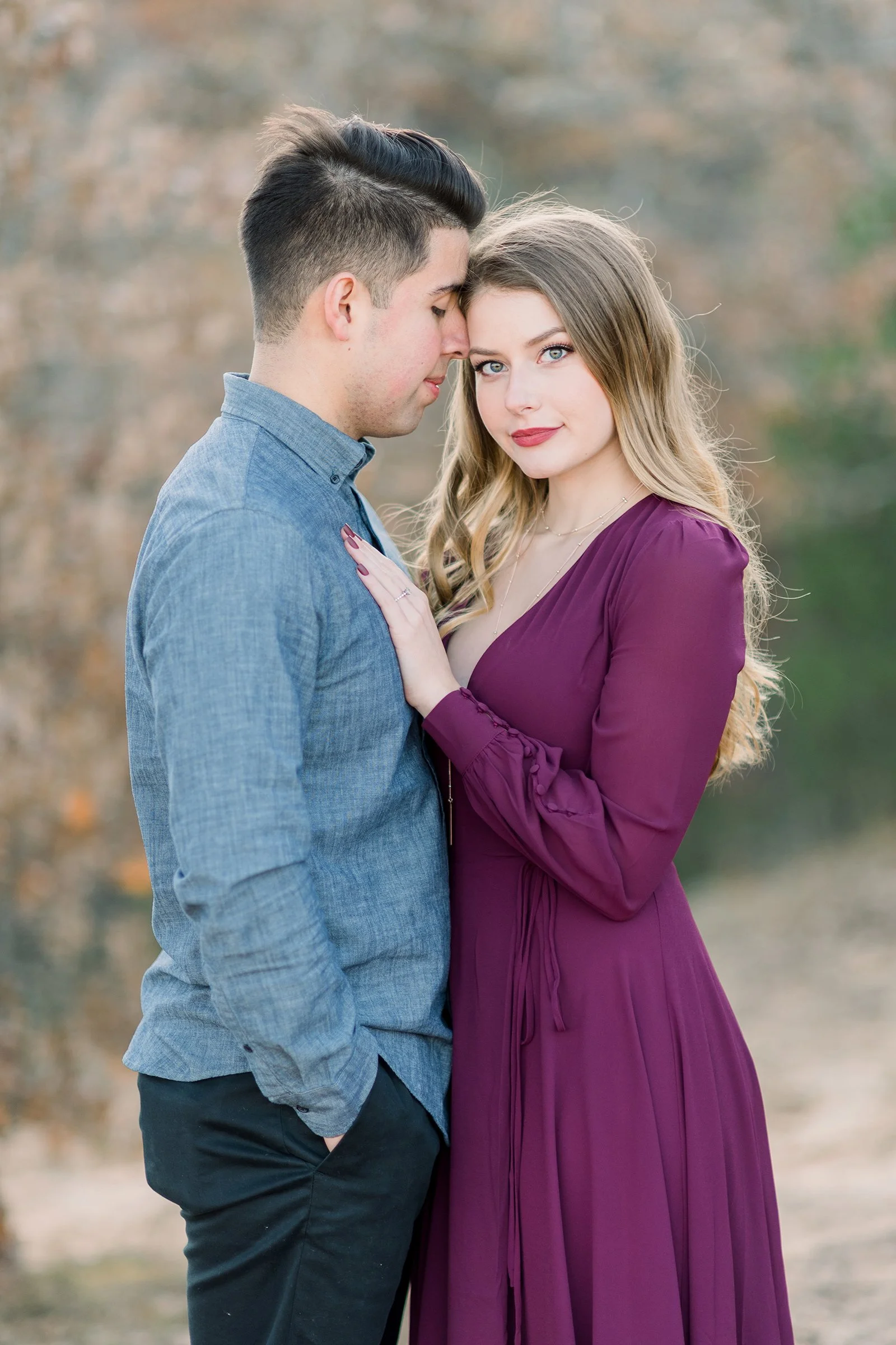 A couple standing close together outdoors during autumn, with the man in a blue shirt and the woman in a purple dress, touching foreheads and gazing at the camera.