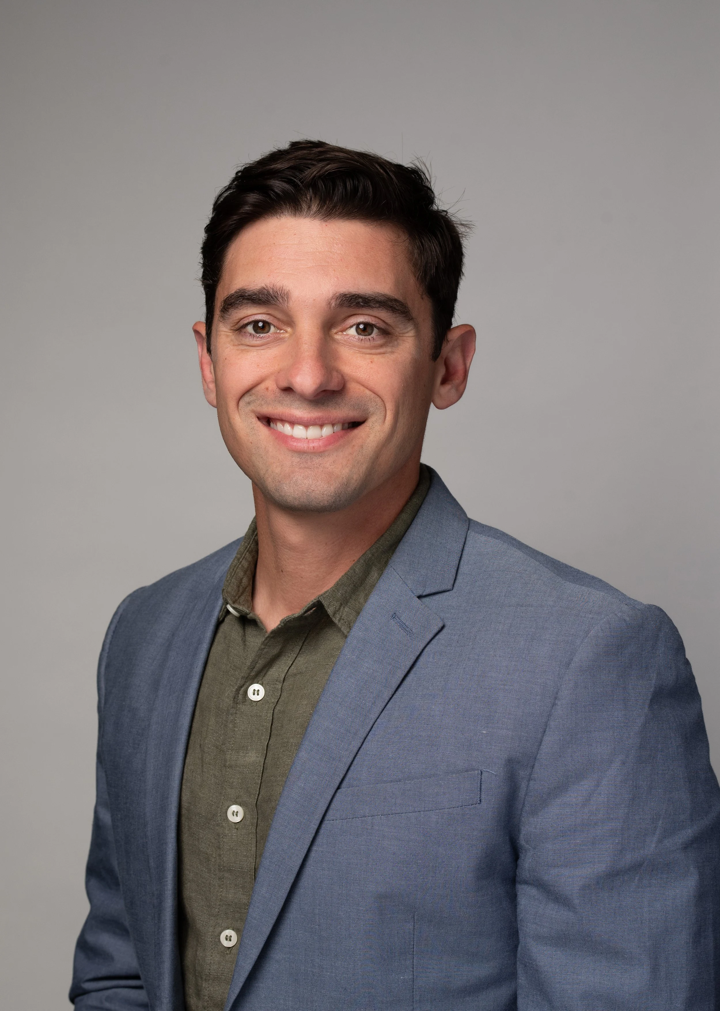 Headshot of a young man with dark hair, wearing a light blue blazer and an olive green shirt, smiling against a neutral background.