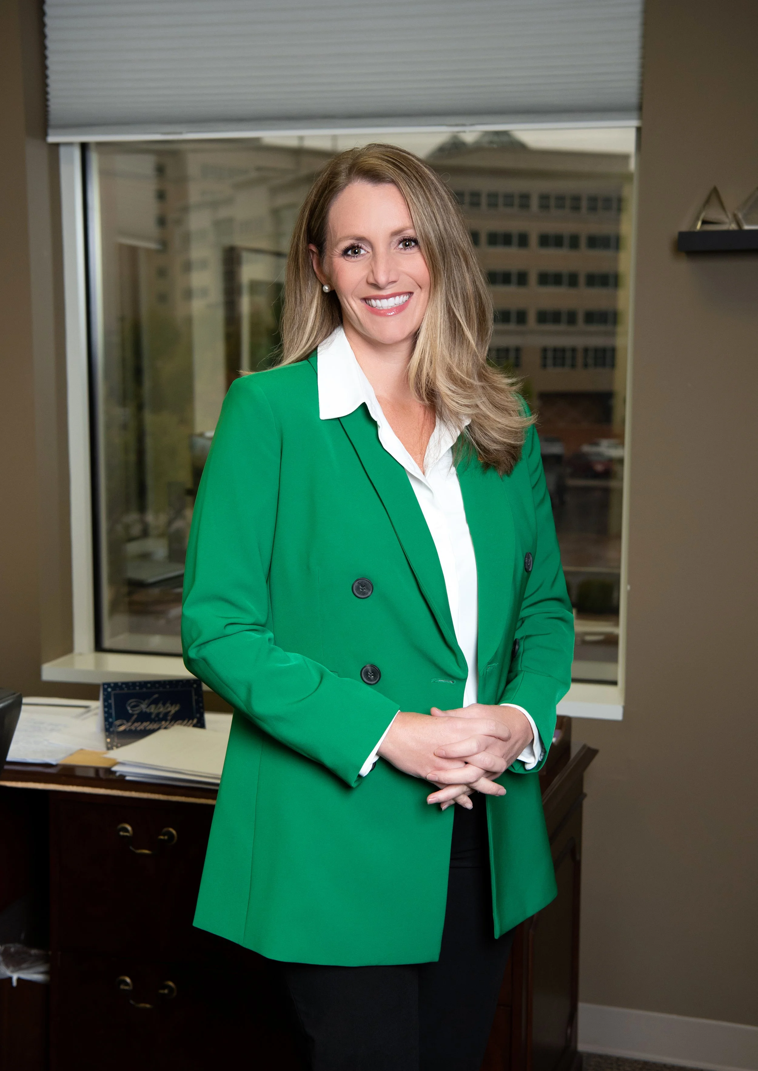 Woman with blonde hair wearing a green blazer and white blouse, standing in an office with a window and city buildings in the background.