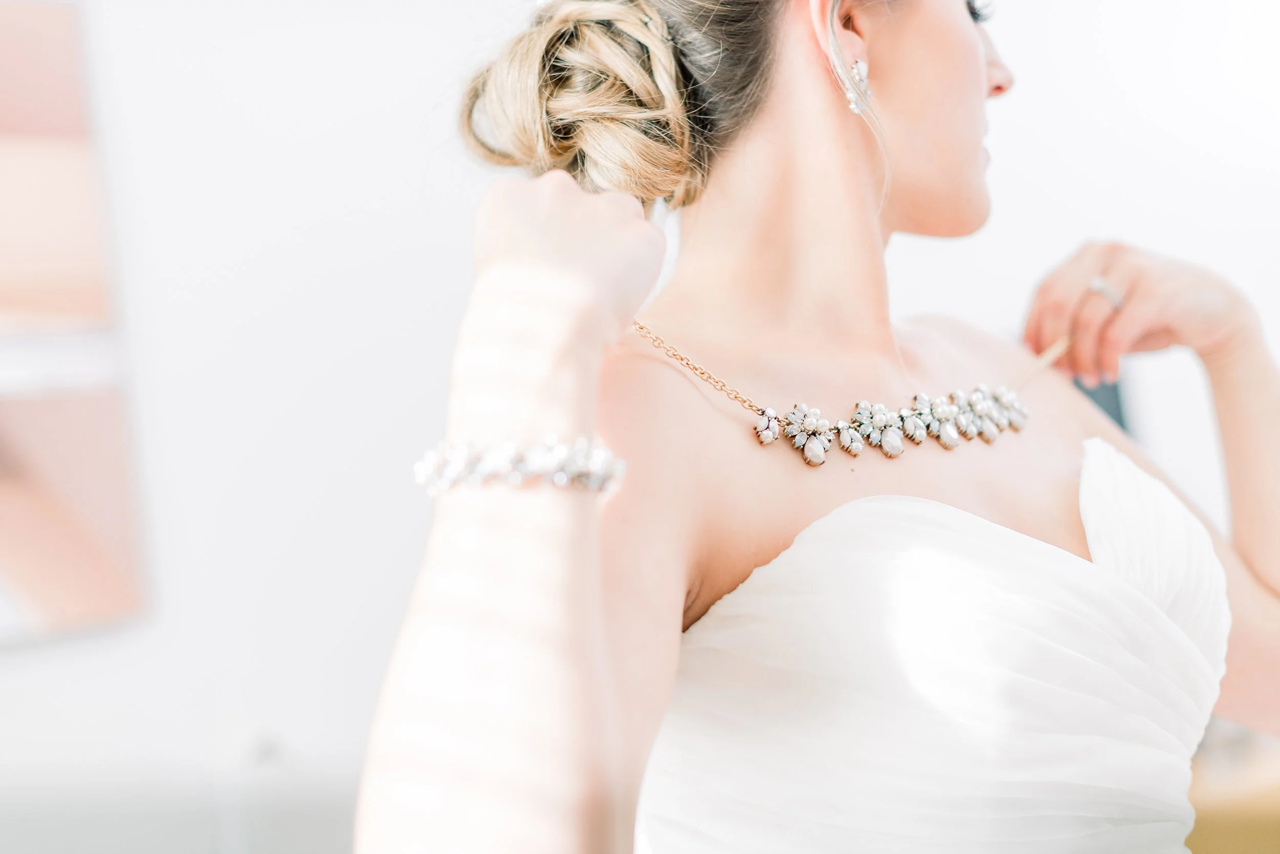 A bride wearing a strapless white wedding dress, adjusting her pearl and rhinestone jewelry, including a necklace, bracelet, and earrings.