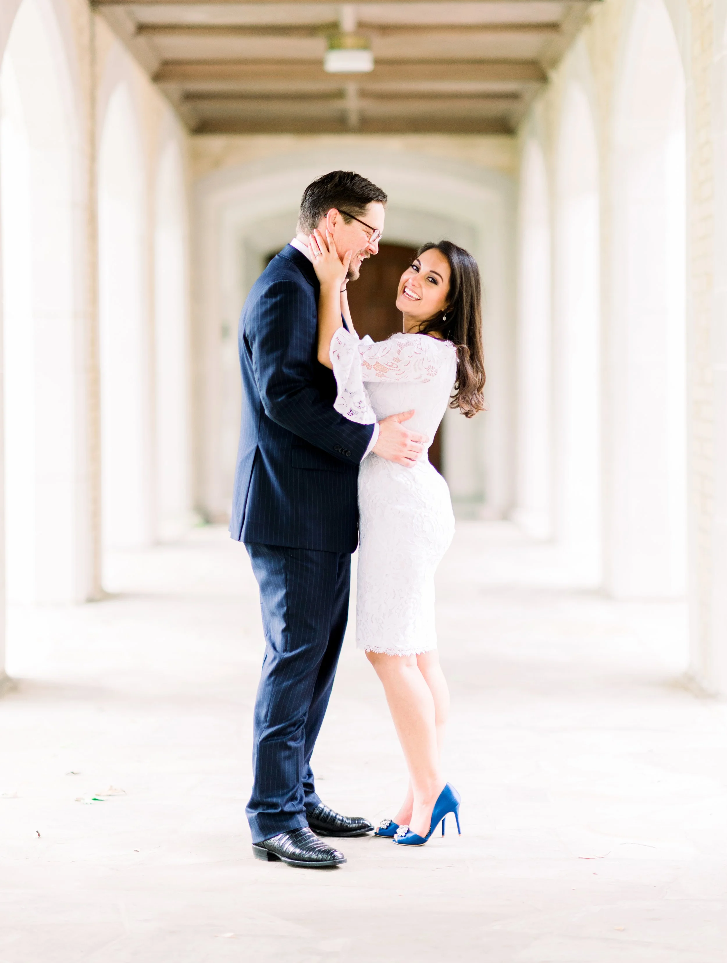 A happy couple in wedding attire standing in a bright corridor, embracing and gazing at each other.