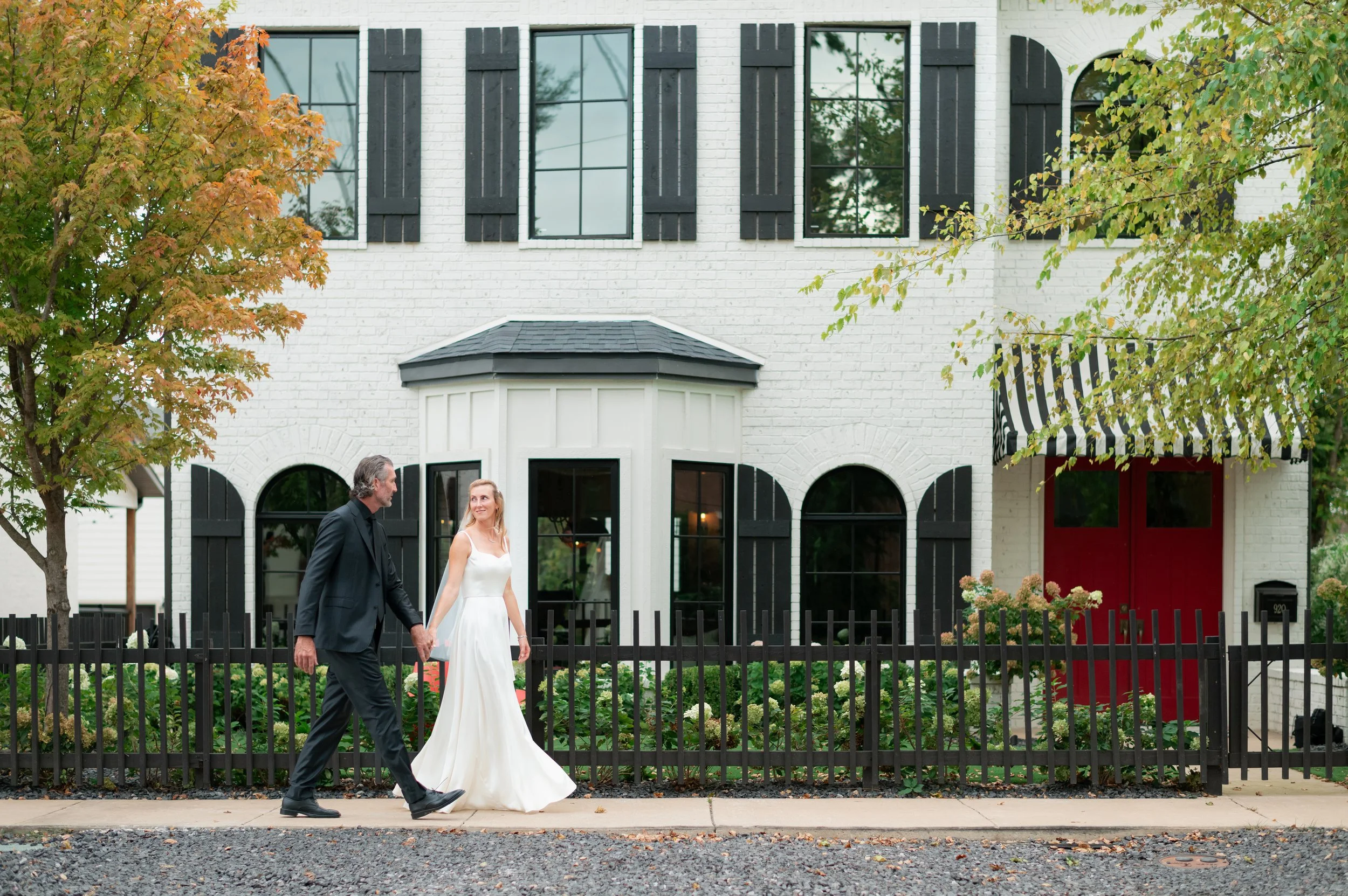A man in a black suit and a woman in a white wedding dress walking hand in hand on a sidewalk in front of a white house with black accents.