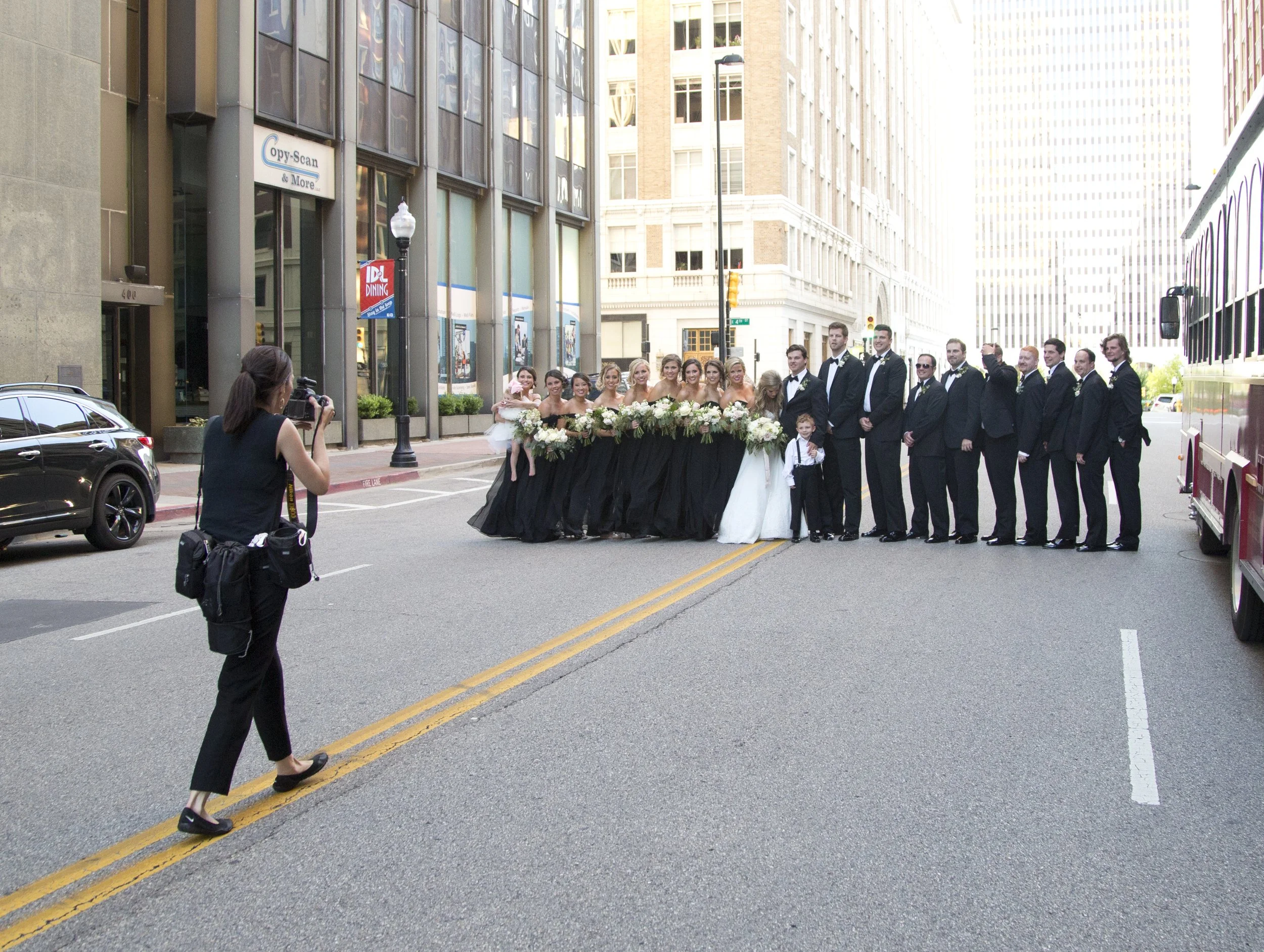A wedding party posing on a city street with a photographer taking their picture, all dressed in formal attire.