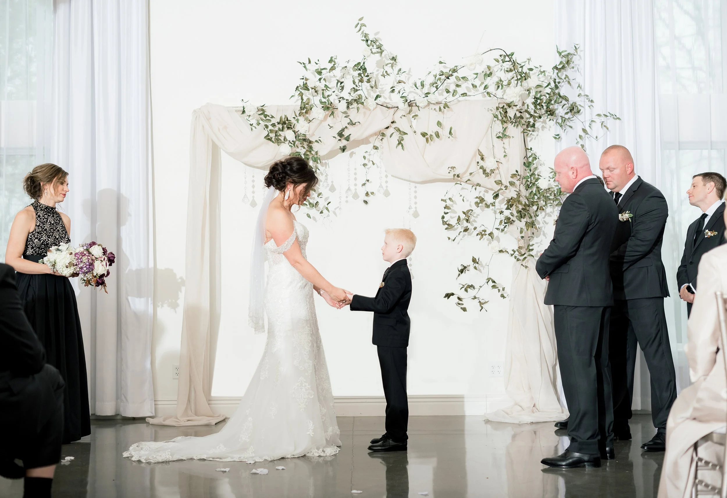 Bride and young boy holding hands during wedding ceremony, surrounded by bridesmaids and groomsmen, in a bright room with floral decorations.