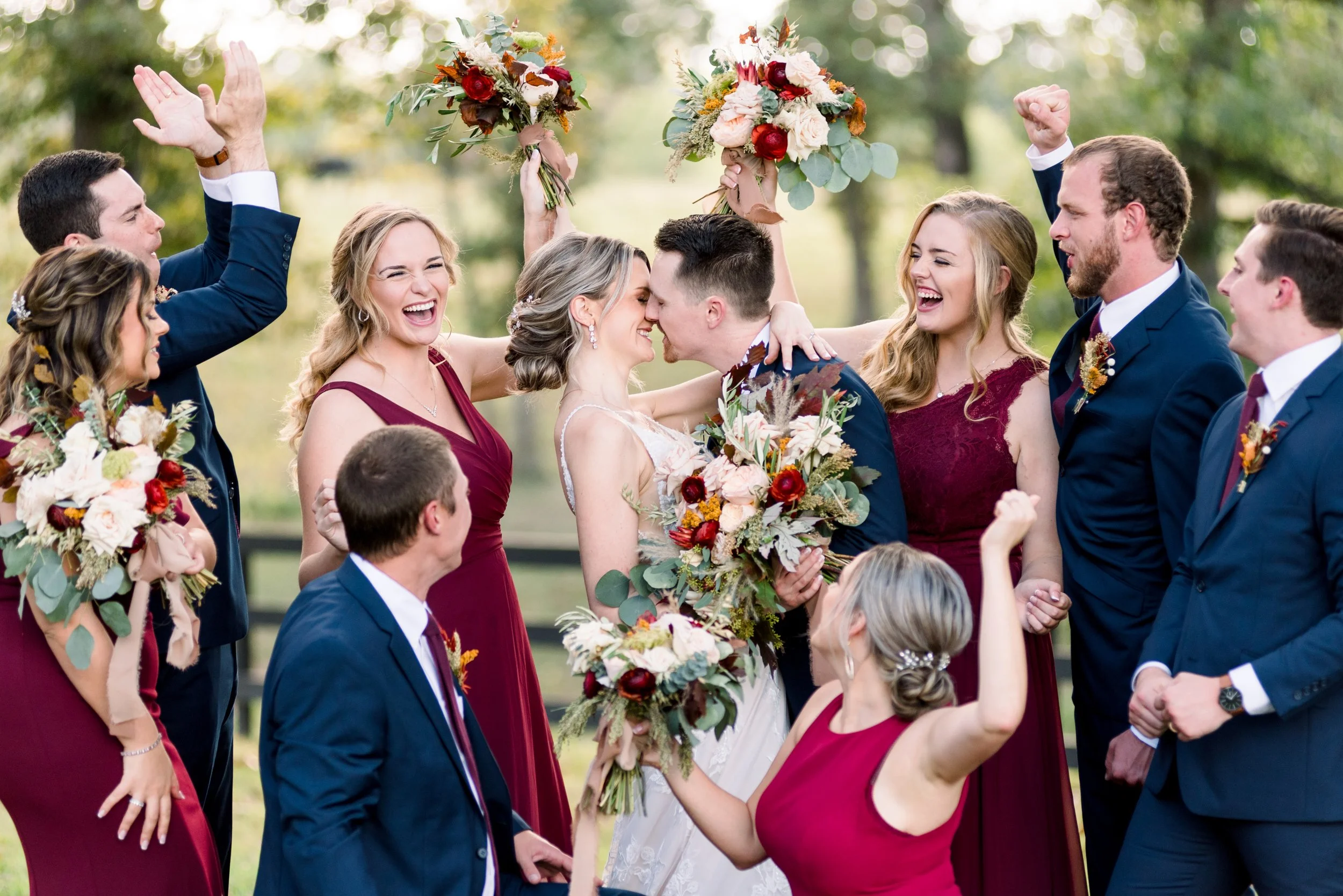 A group of people celebrating a wedding outdoors, with the bride and groom in the center. The guests are joyfully cheering and holding bouquets, dressed in formal attire, with a background of trees and sunlight.
