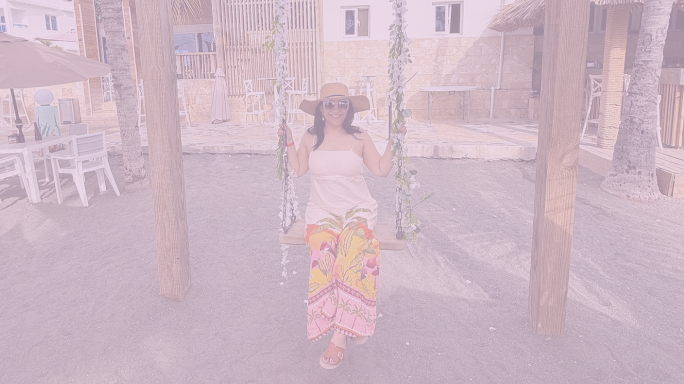 Mujer con sombrero grande y gafas de sol sentada en un columpio con decoración de flores en un entorno exterior con mesas y sillas blancas.