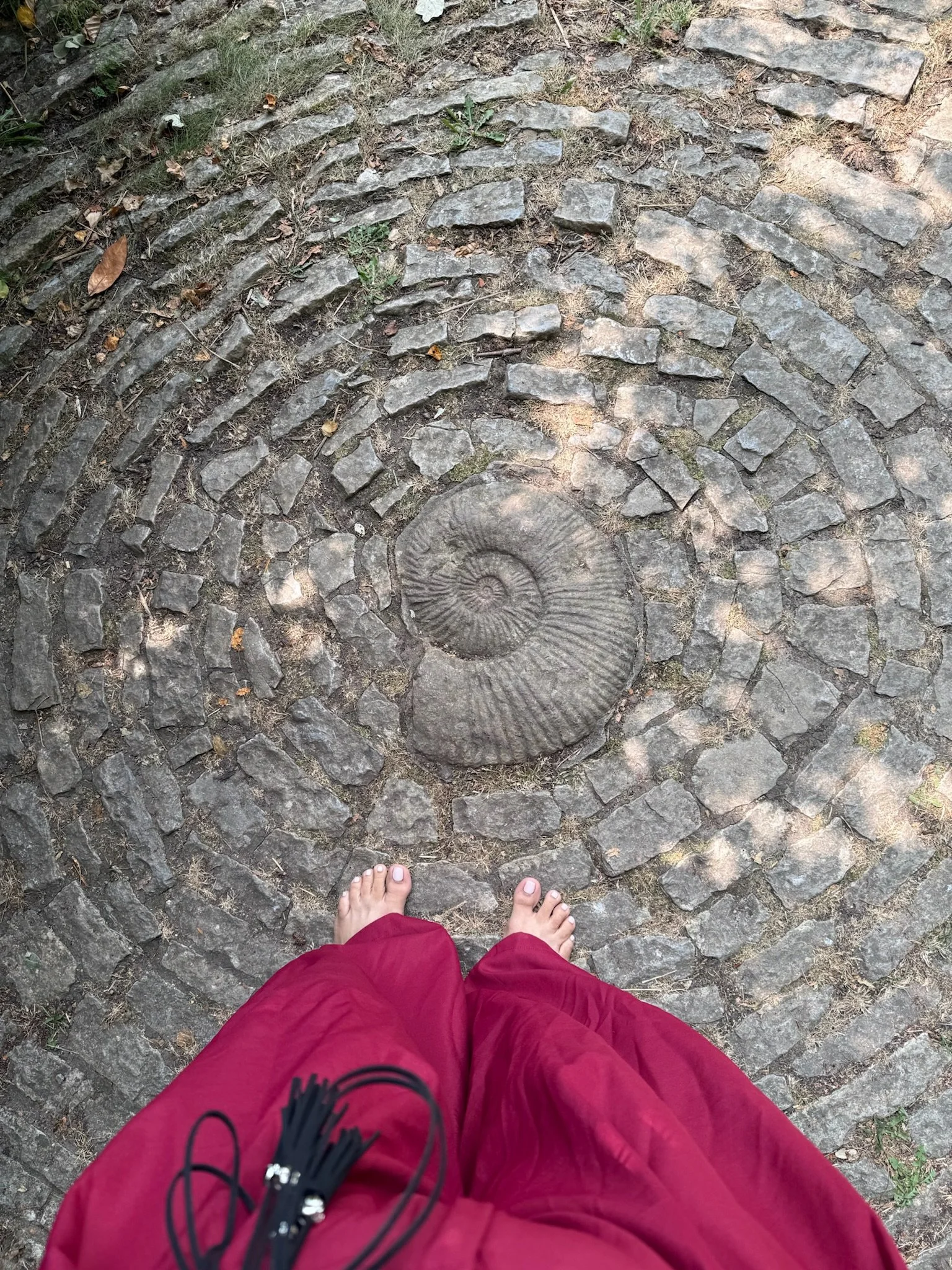 Vista desde arriba de una persona con pantalones rojos y camiseta negra, de pie sobre un camino de piedras en espiral, con pies descalzos, rodeado de tierra y hojas secas.