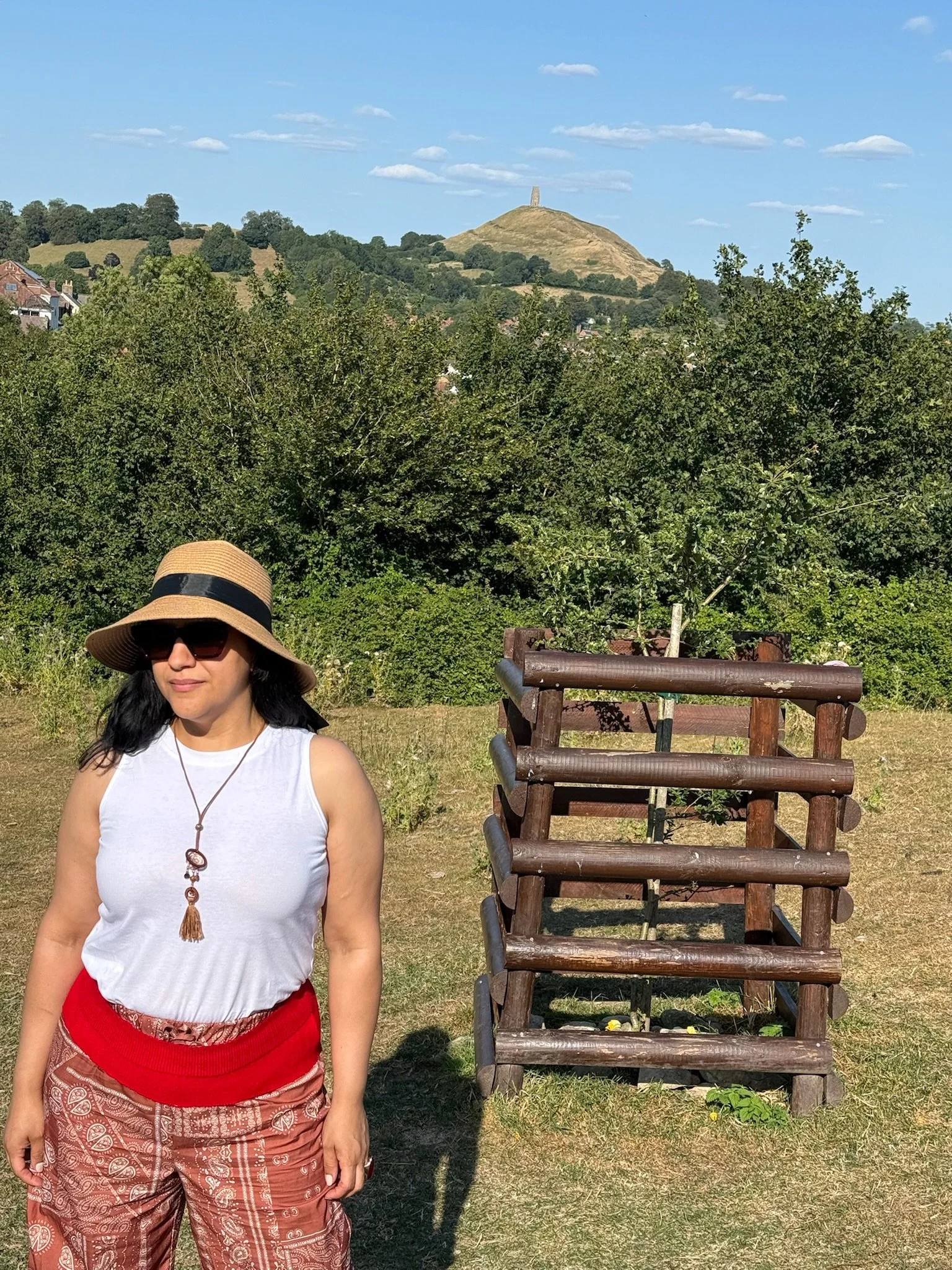 Mujer con sombrero, gafas y ropa blanca y roja, en un área al aire libre con árboles y un cerro en el fondo, en un día soleado y despejado.